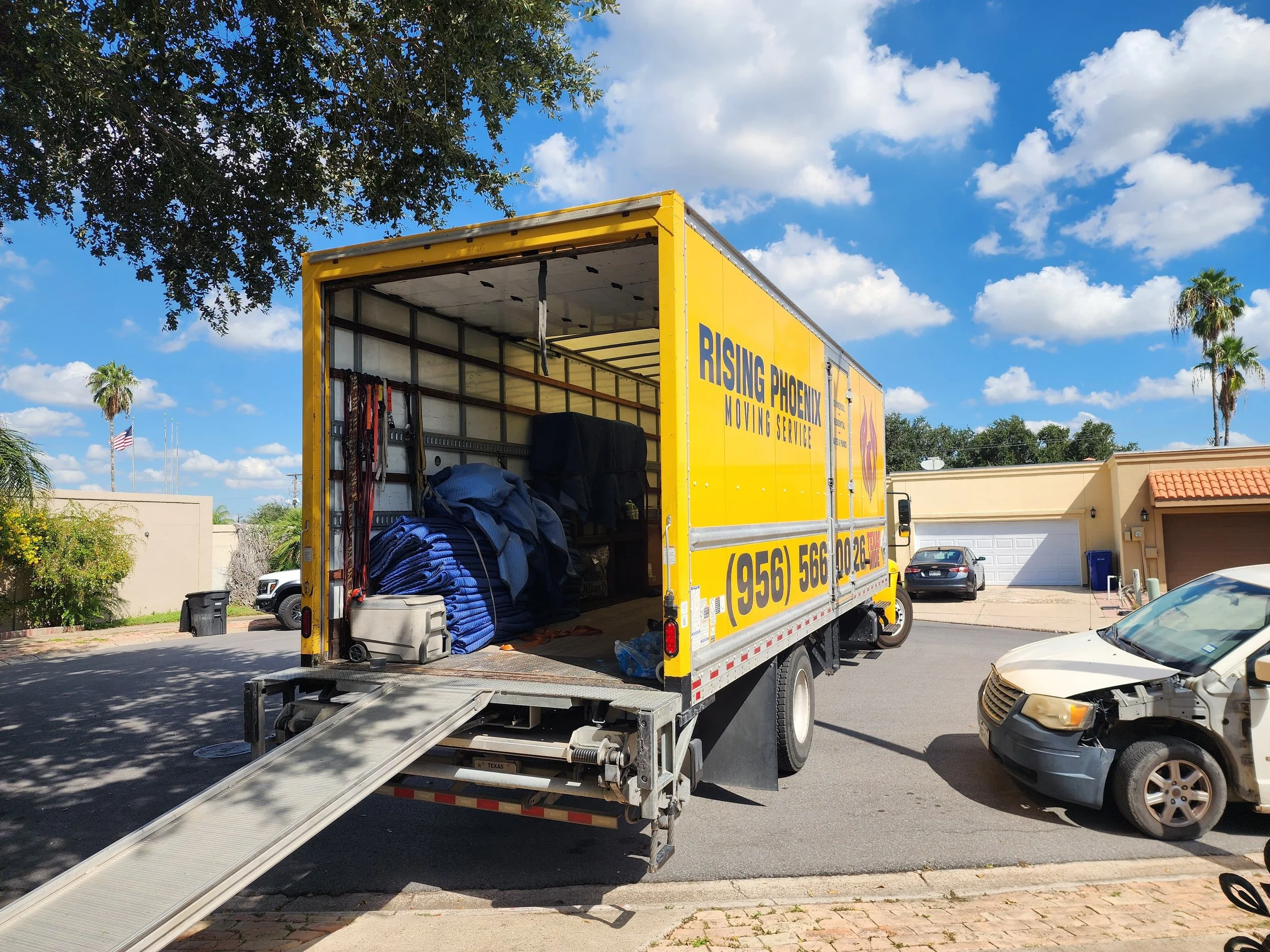 A yellow moving truck labeled 'Rising Phoenix Moving Service' with an open back filled with moving supplies, parked in a residential neighborhood with a sunny sky, some clouds, and trees.