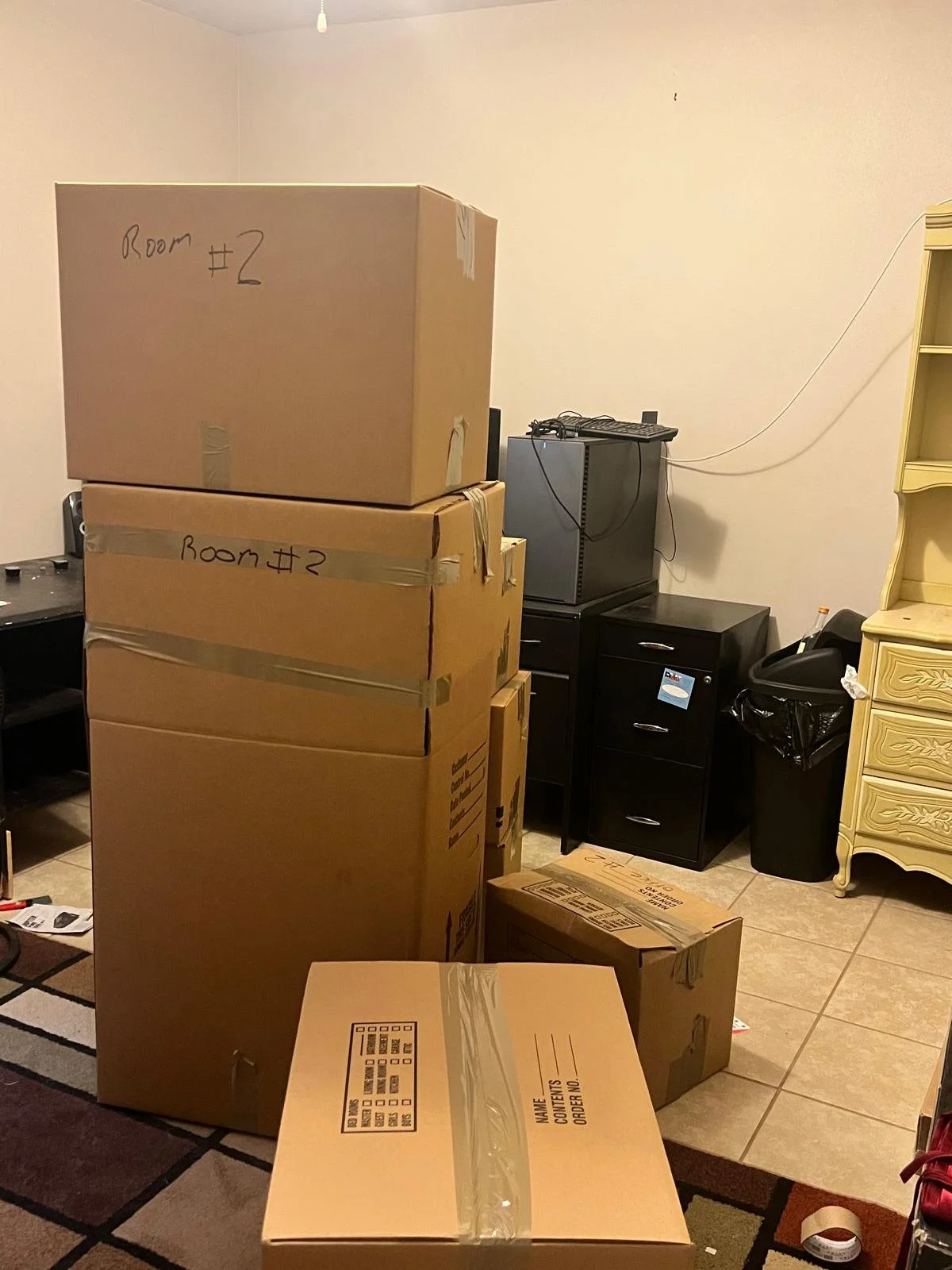 Rising Phoenix Moving Service team packed the room filled with moving boxes labeled 'Room #2,' stacked in front of a black desk with a small TV on top, a black dresser, and a yellow dresser, with cords visible on the wall.