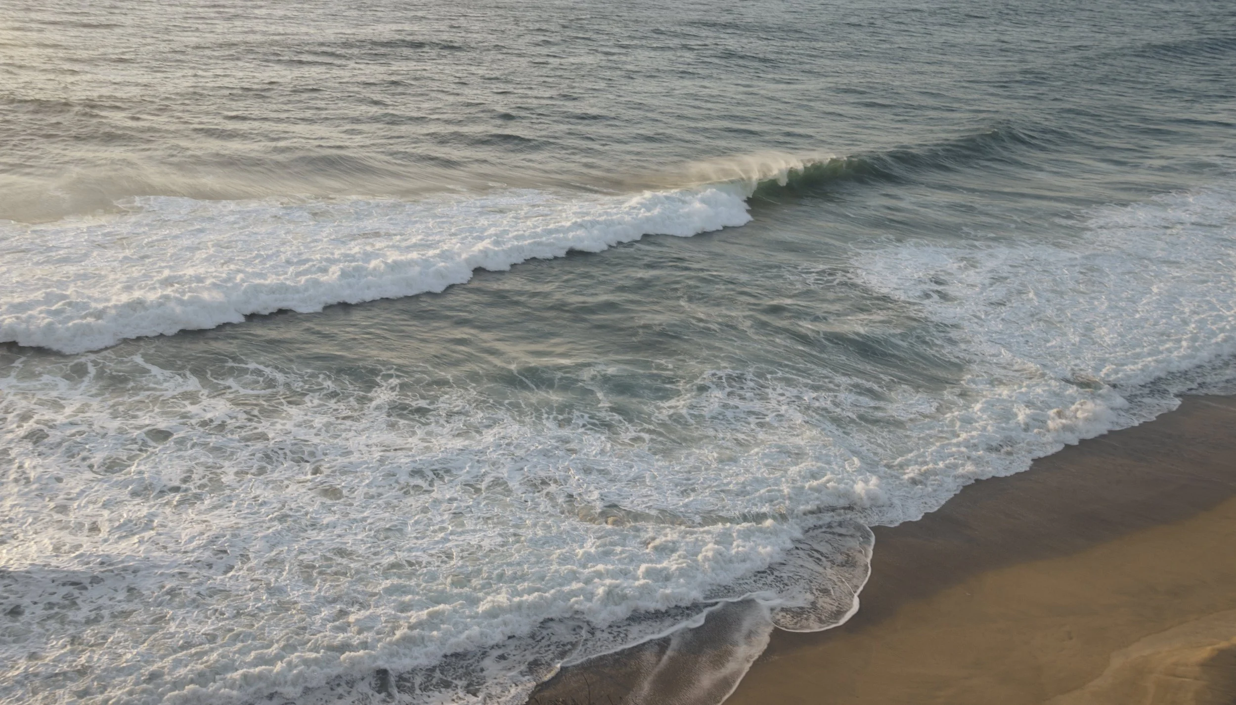 Aerial view of the ocean with waves breaking on the sandy beach at sunset.