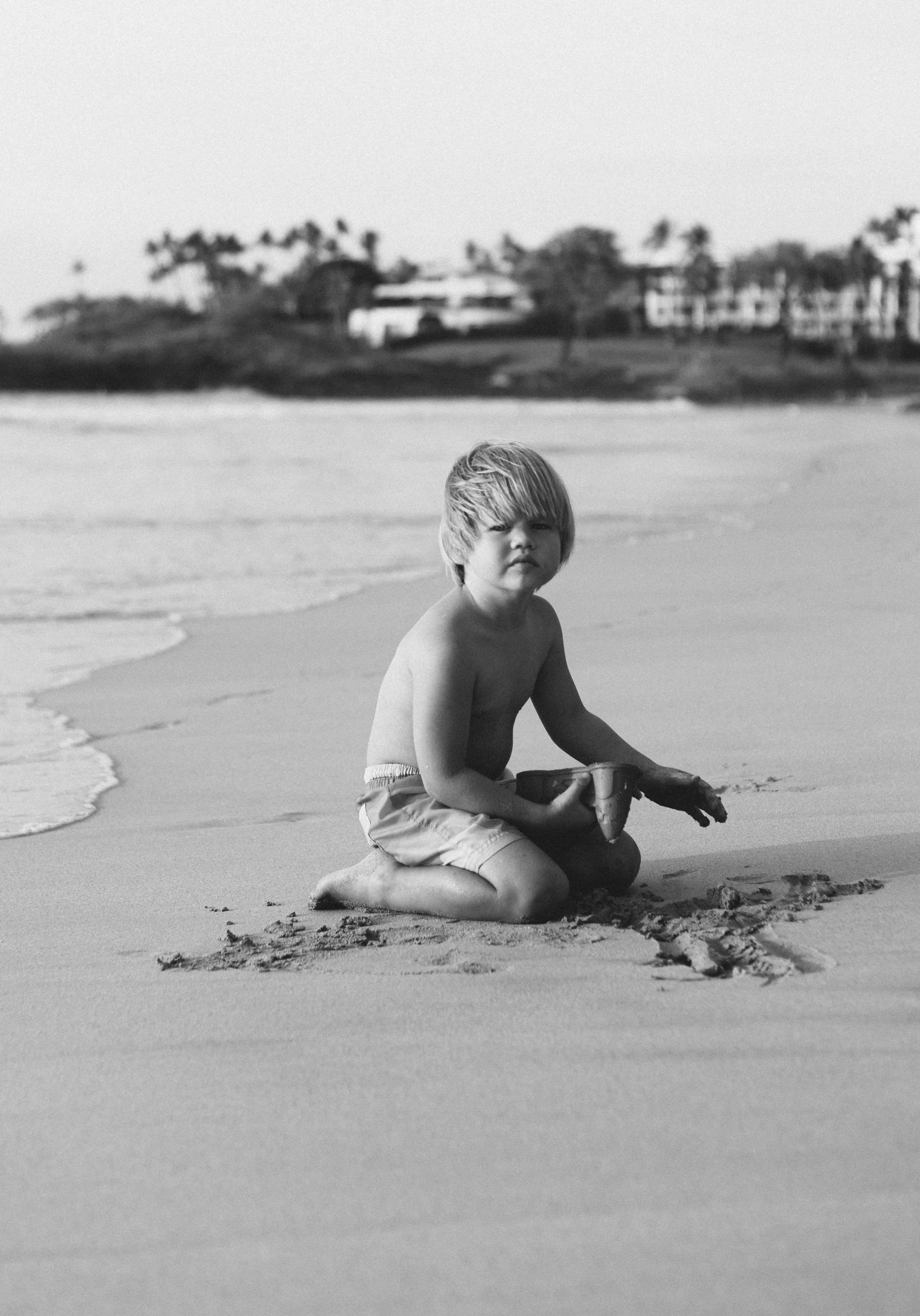 A young boy with messy hair, sitting on sand at the beach, holding a toy fish, with shoreline and distant trees and buildings in the background, in black and white.