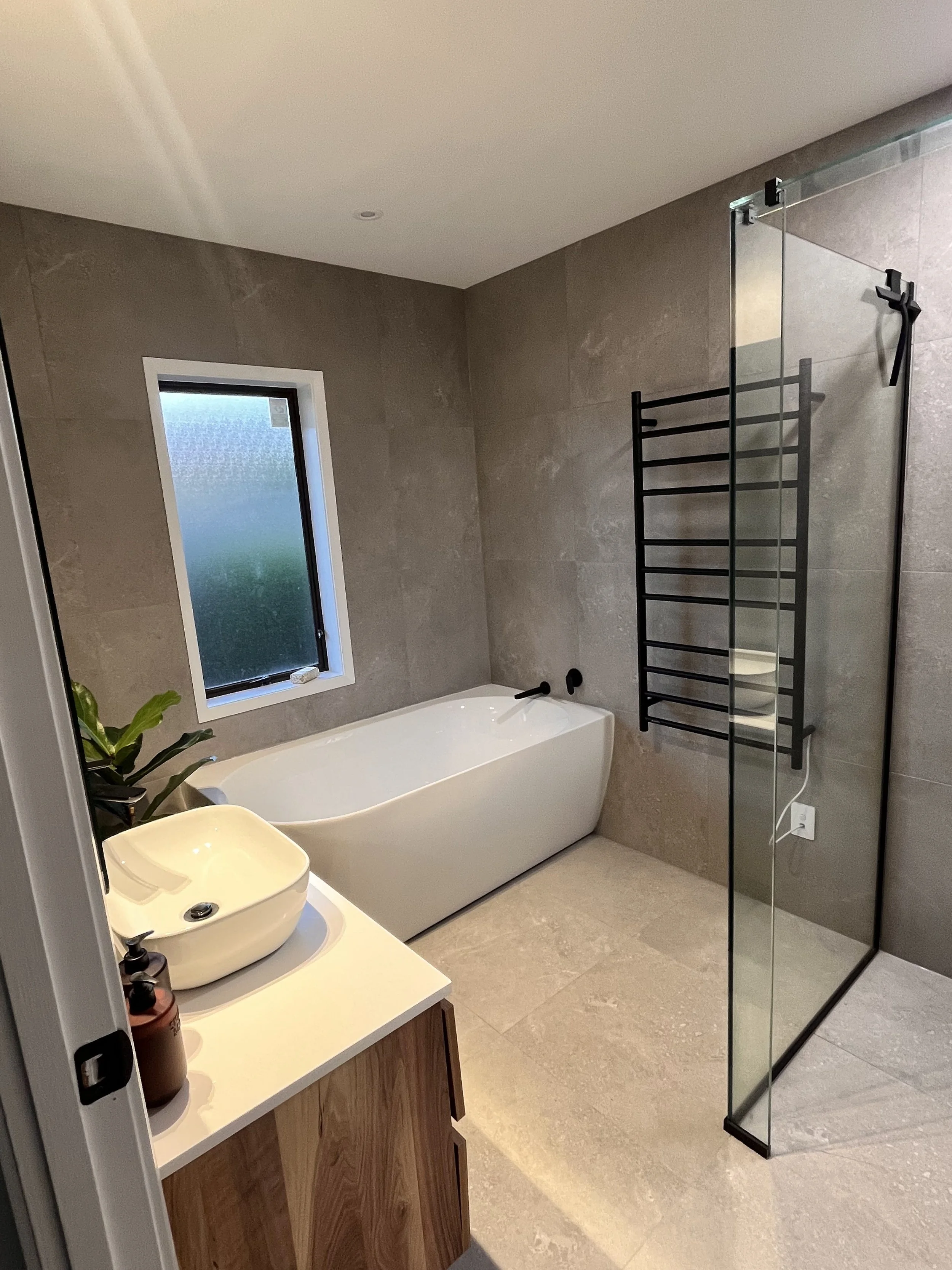 Modern bathroom with a freestanding bathtub, glass shower enclosure, vessel sink on a wooden vanity, tiled walls and floor, wall-mounted towel rack, and a small plant.