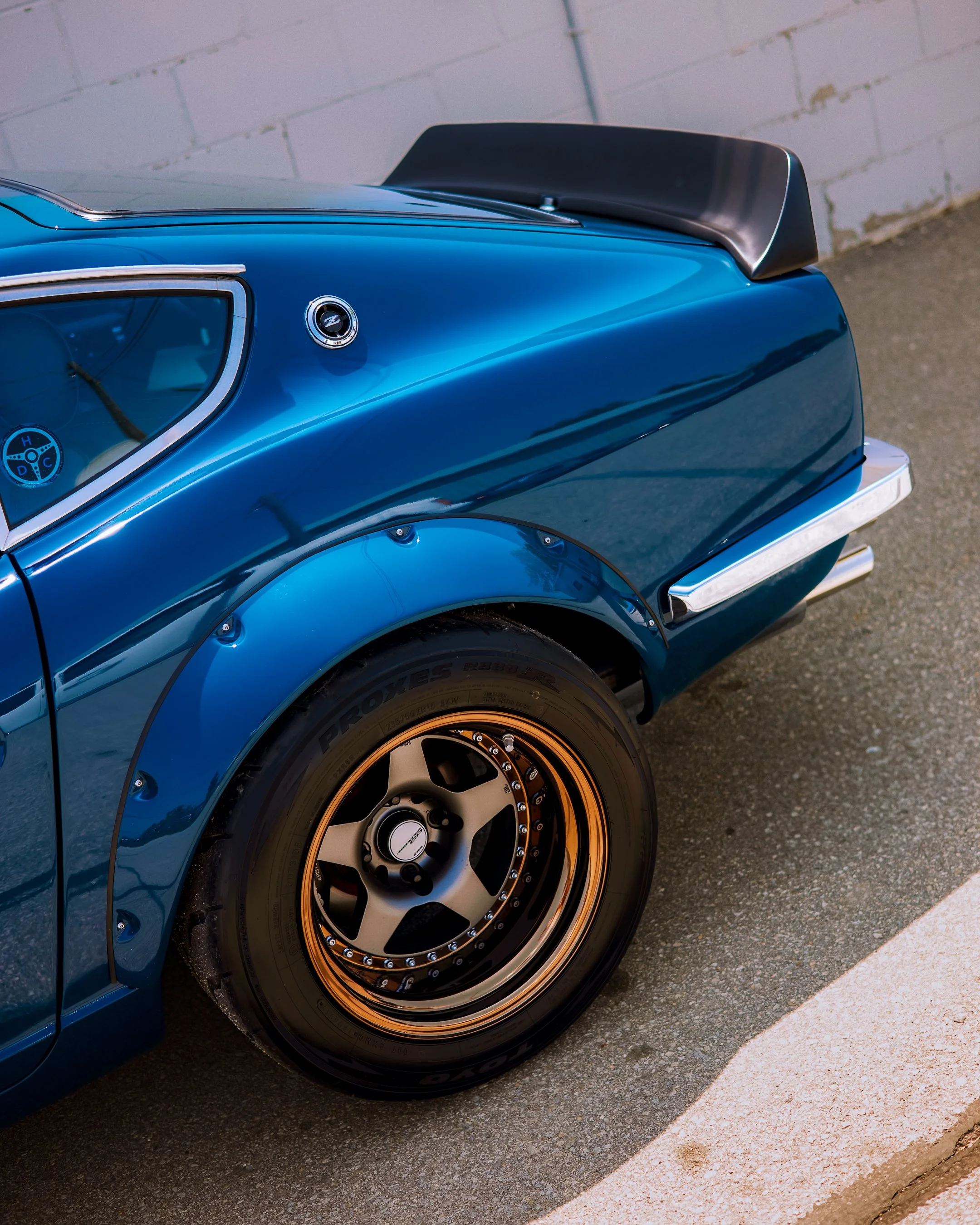 Close-up of the rear left side of a classic blue car with a black spoiler, showcasing a gold and black wheel and tire, and part of the rear fender and window.