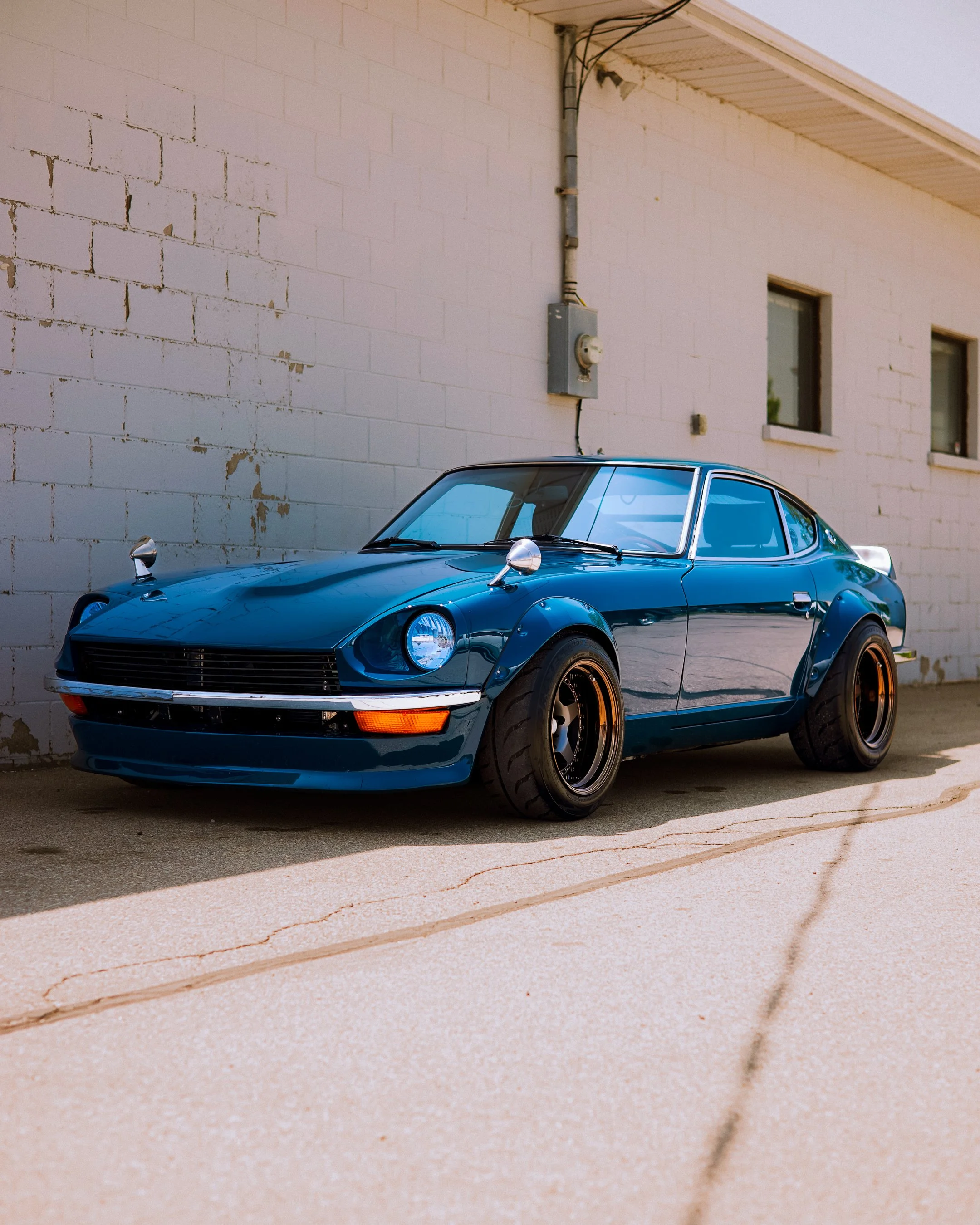 A vintage blue sports car with wide black tires parked against a white brick wall with windows.