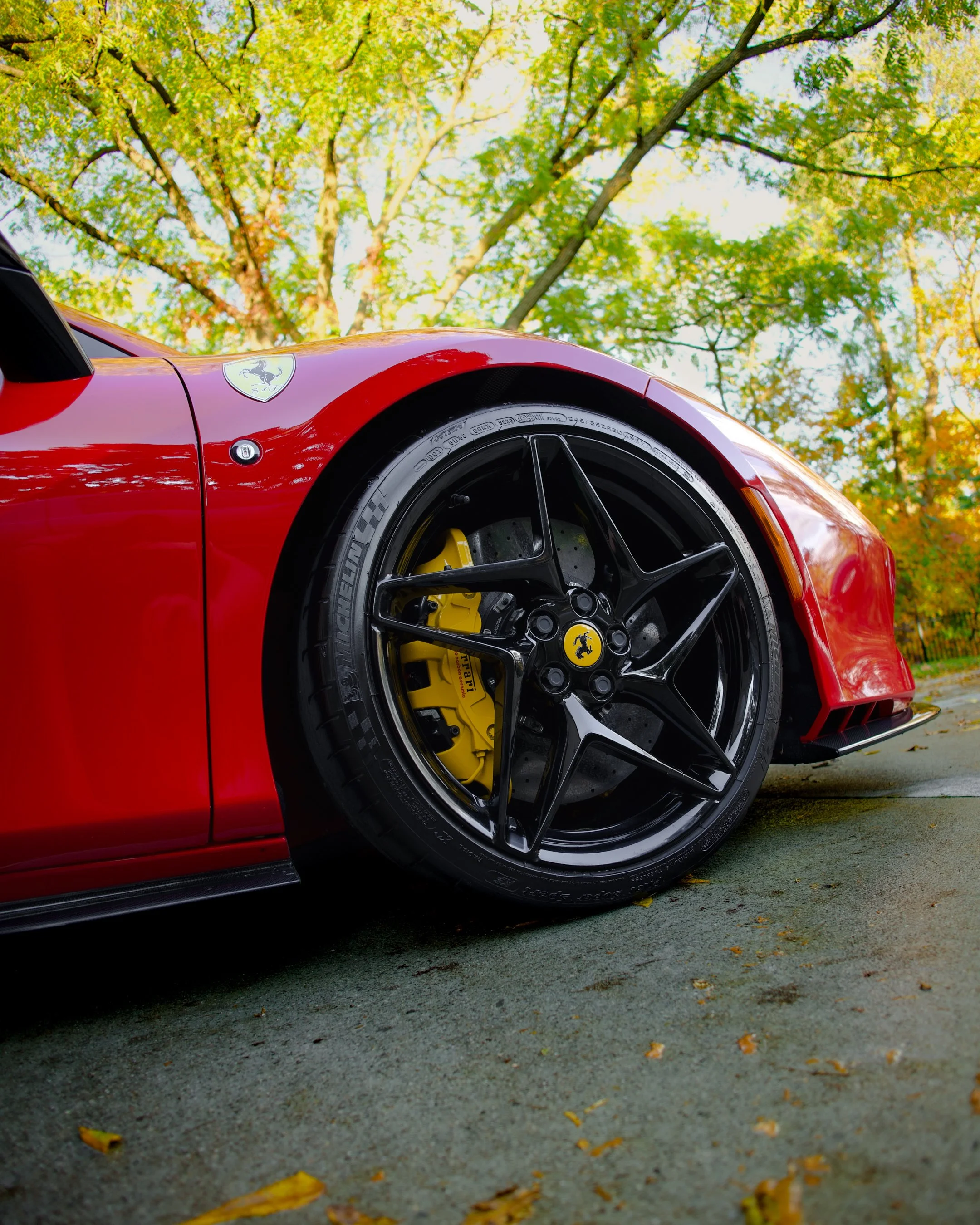 Close-up of a red Ferrari sports car's front wheel and fender, showing black alloy rims, yellow brake calipers, and labeled tires, with green trees and autumn leaves in the background.
