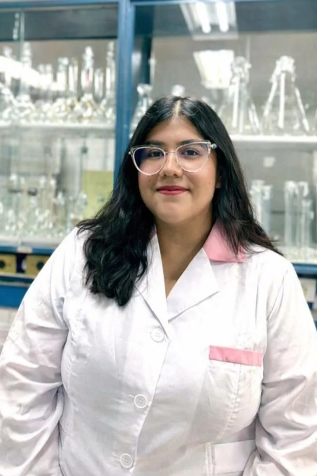 Person smiling indoors with long hair, wearing a 'Friday the 13th' T-shirt and a lanyard, seated at a counter with shelves in the background.