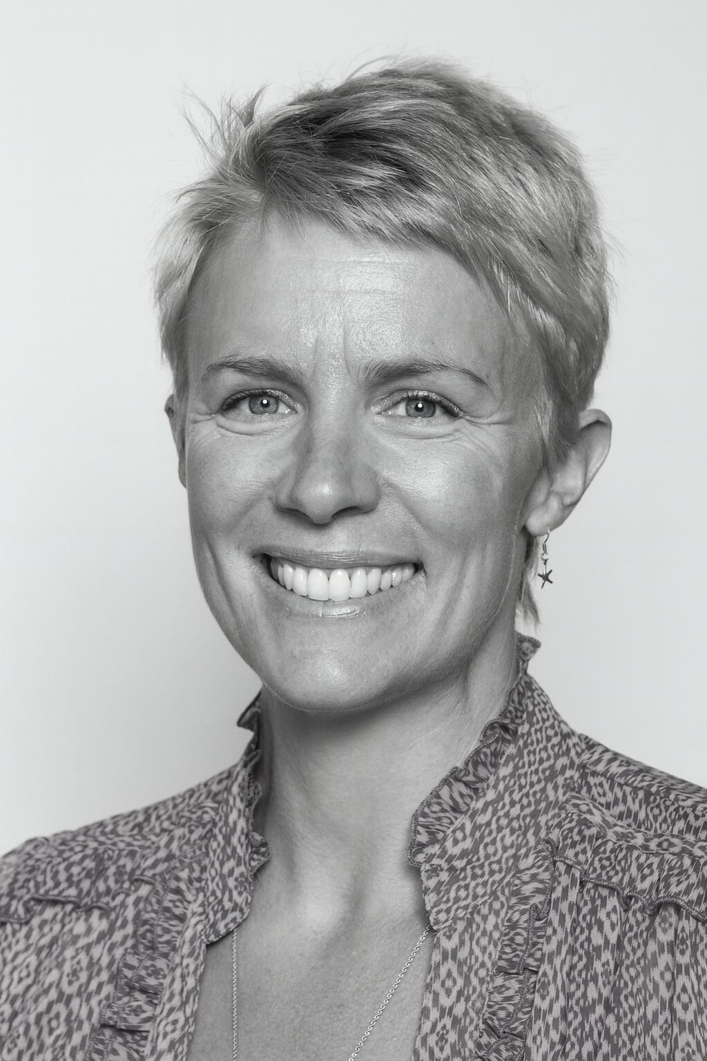 A photo of Victoria Schmidt, A Black-and-white portrait of a smiling woman with short, tousled hair, wearing a patterned blouse and star-shaped earring.