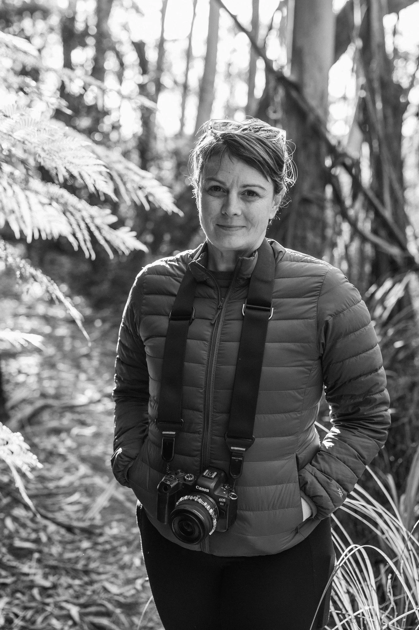 A photo of Karen Webb, a woman stands in a forested area with ferns and trees in the background. She is wearing a puffy jacket, has a camera hanging around her neck, and has sunglasses resting on her head.
