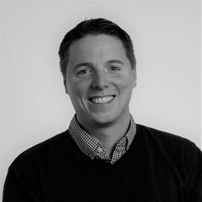 A photo of Arno Parolini, a black and white portrait of a smiling man wearing a checkered shirt and a dark sweater.