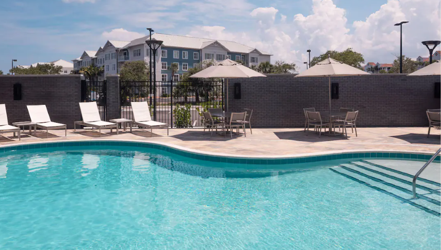 Swimming pool area with lounge chairs, tables, umbrellas, and a brick wall, with residential buildings in the background under a partly cloudy sky.