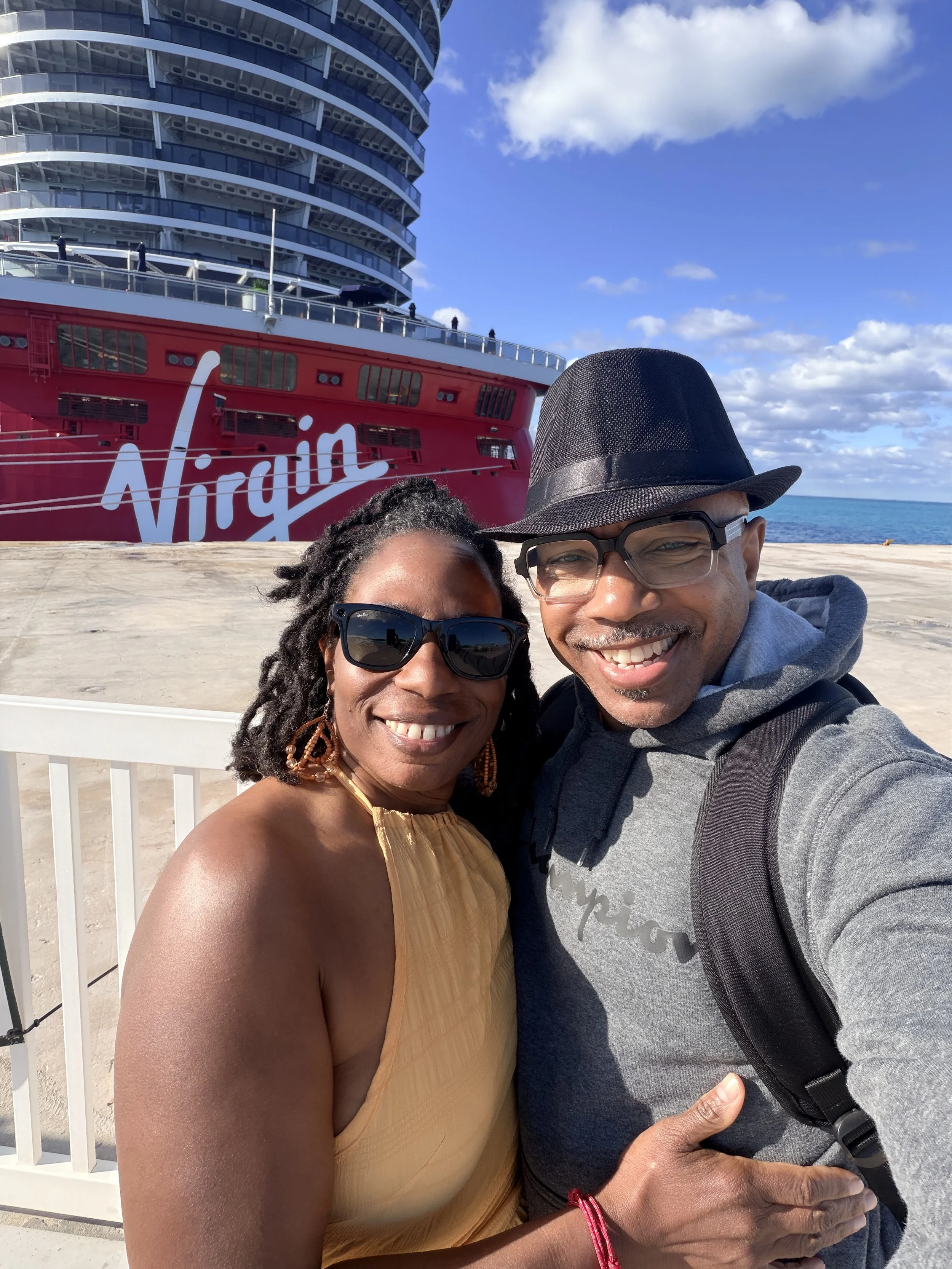 A happy couple taking a selfie in front of a Virgin Voyages cruise ship. Couple enjoying Virgin Voyages cruise experience during a romantic getaway.