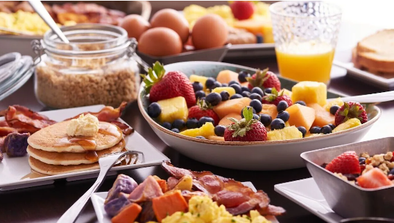 A breakfast spread including a bowl of mixed berries and pineapple, scrambled eggs, bacon, pancakes with butter, a jar of brown sugar, eggs, orange juice, and various other breakfast foods.