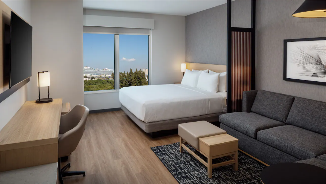 Hotel room with a large window, bed, gray sofa, wooden desk with a monitor, chair, and a black and white wall art.