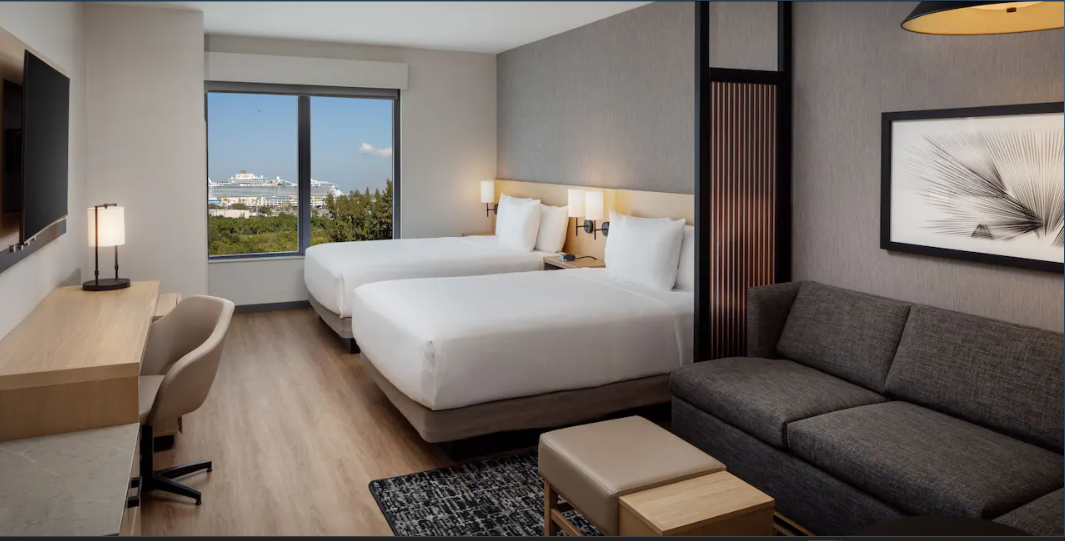 Hotel room with two queen beds, a sofa, a desk, and a window showing a view of a cruise ship and greenery.