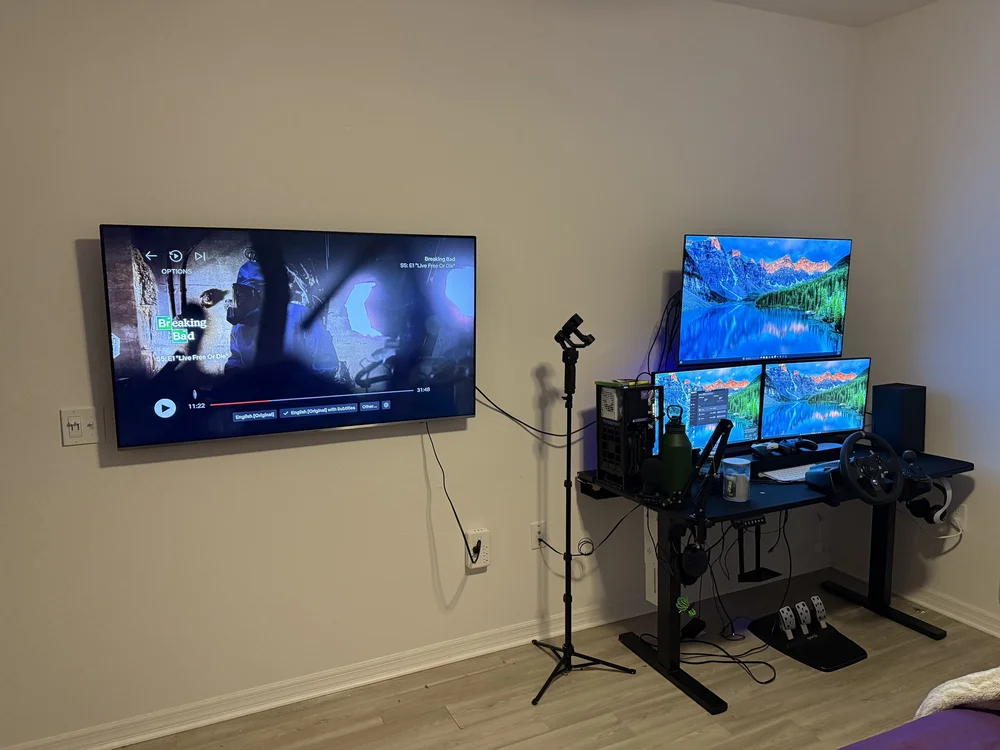 Wall-mounted TV with gaming setup featuring three monitors and racing wheel