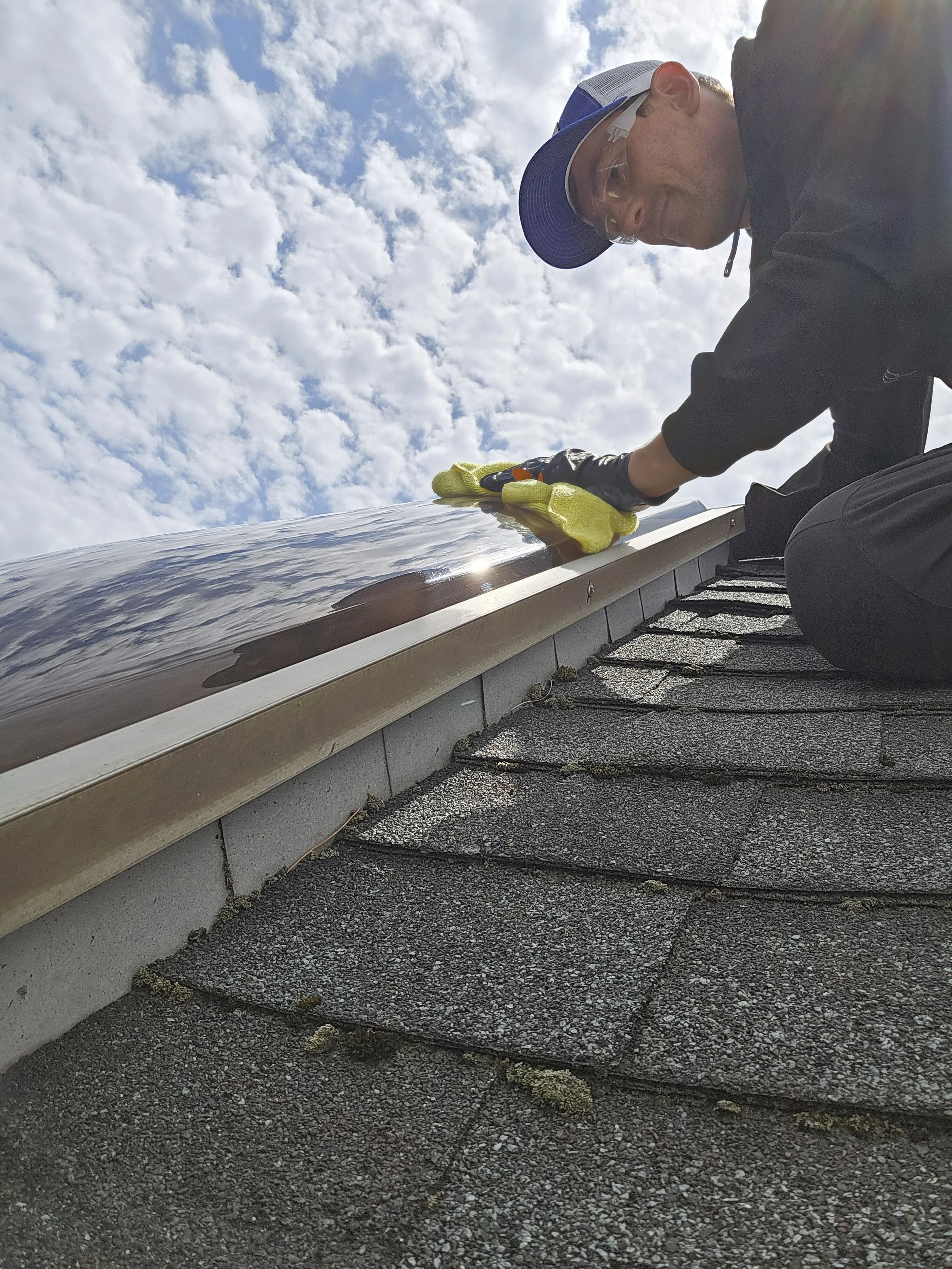 A man working on his roof, cleaning or inspecting the gutter with a cloth and gloves, under a partly cloudy sky.