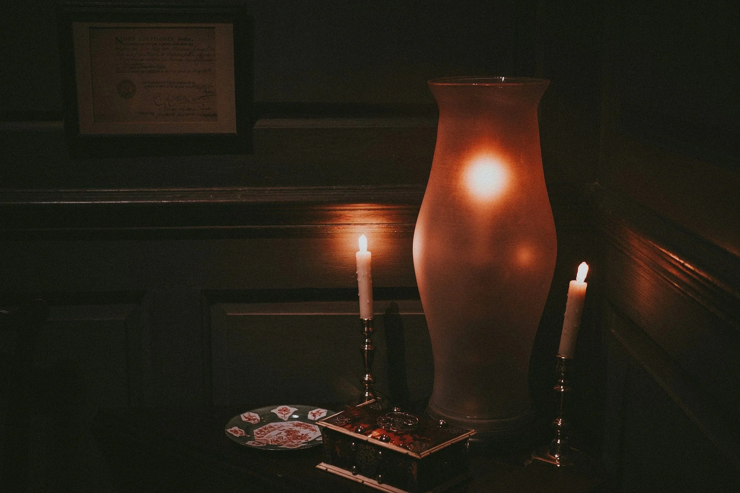 Dimly lit table with a tall, frosted glass vase, two lit candles in silver holders, a decorative plate, and a framed certificate in the background.