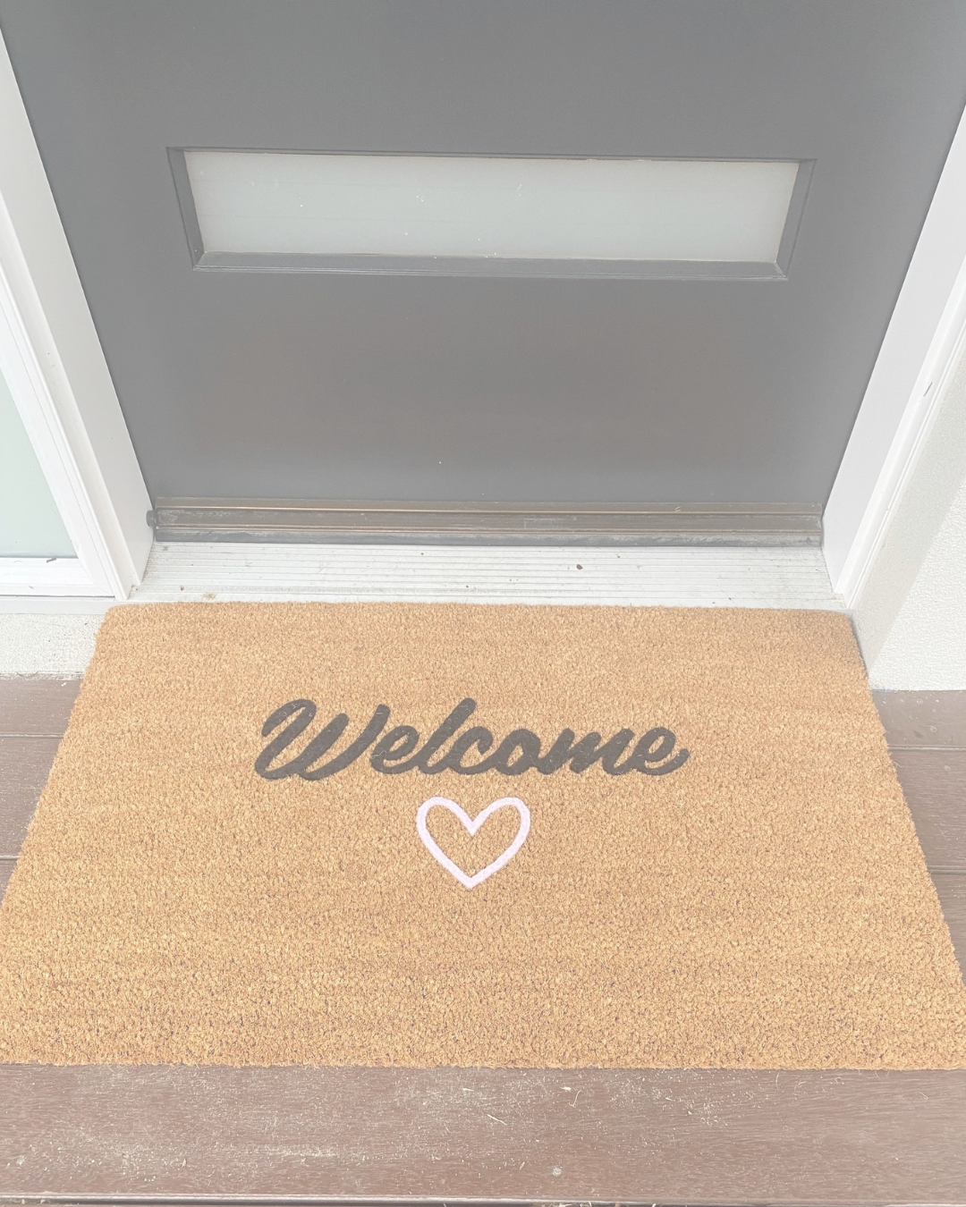 Doormat with 'Welcome' text and heart design in front of a gray door.