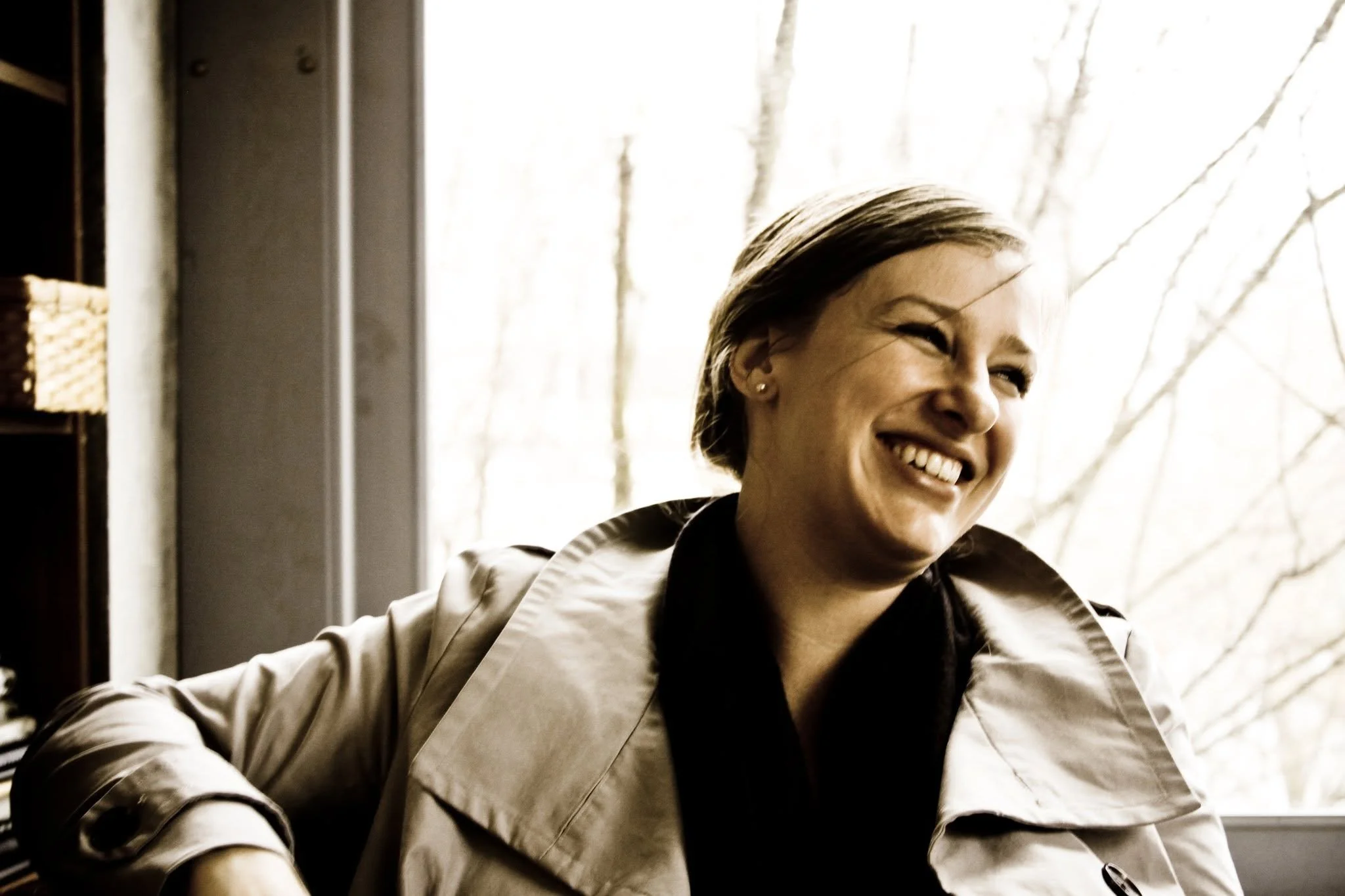 Cristin with her hair pulled back smiling and wearing a beige jacket, sitting indoors near a window with light streaming in, and a bookshelf partially visible on the left side.
