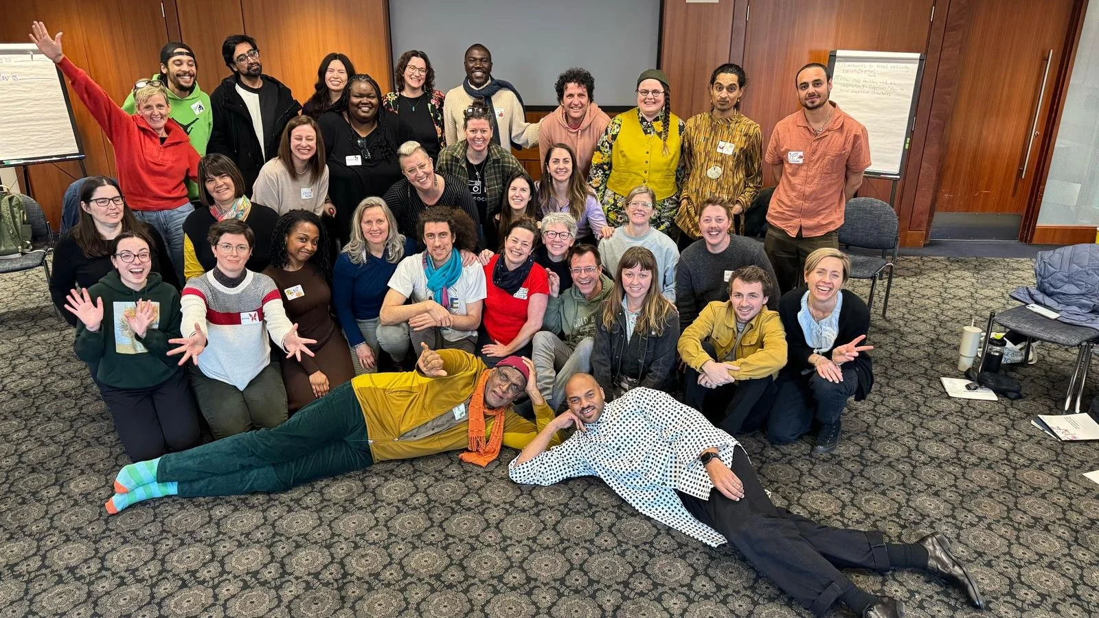 Group of diverse adults in a conference room, smiling and posing for a photo. Some people are sitting on the floor, others are standing, with two individuals lying in front. Background includes flipcharts and wooden paneled walls.