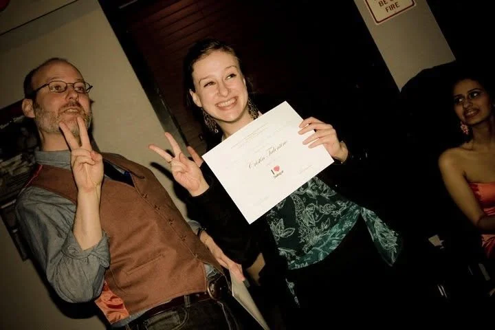 Cristin smiling and holding a certificate, standing next to a man making a peace sign, with another woman seated in the background.