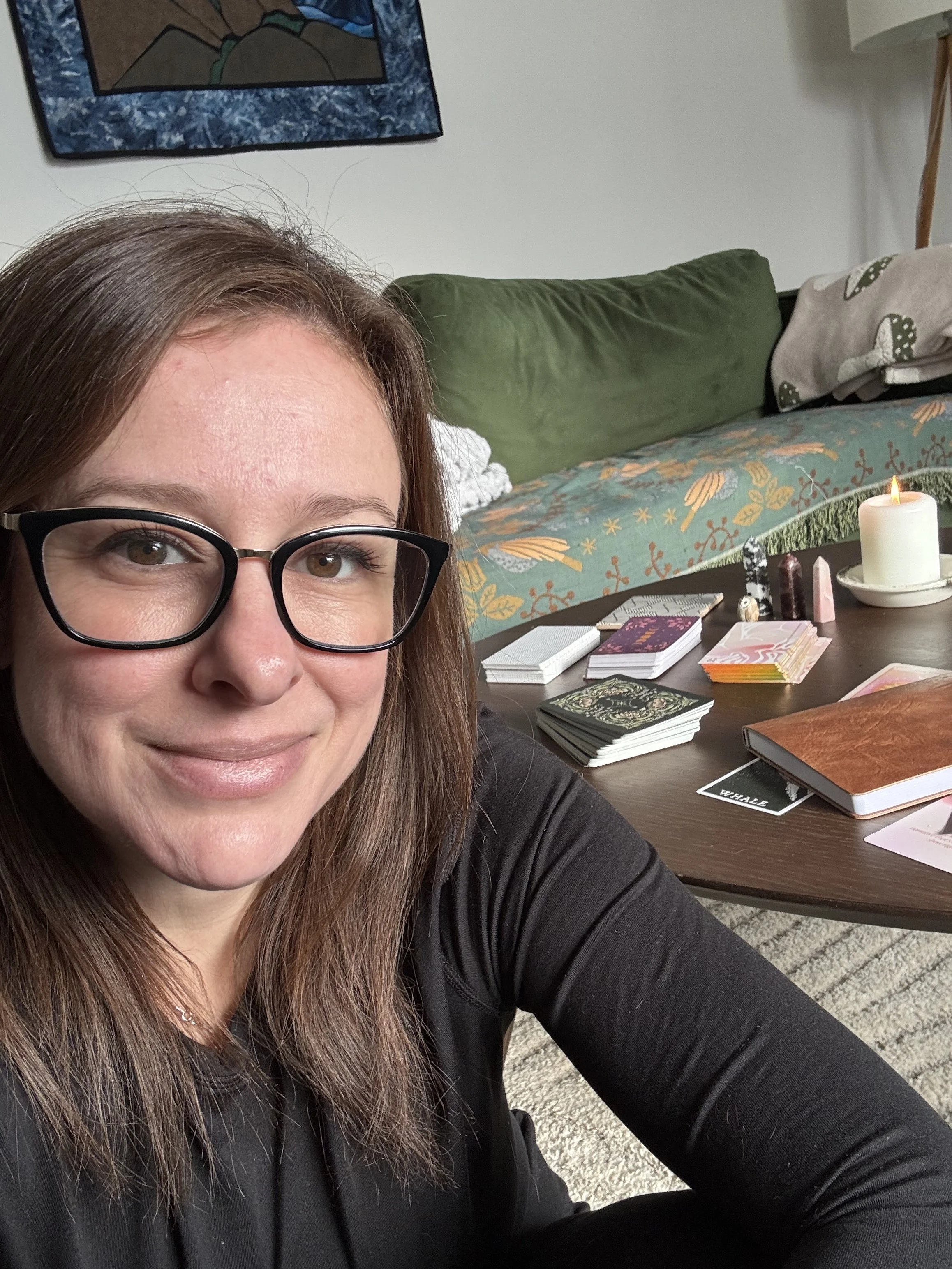 Cristin with glasses taking a selfie indoors, with tarot cards, a candle, and a book on a table in the background.