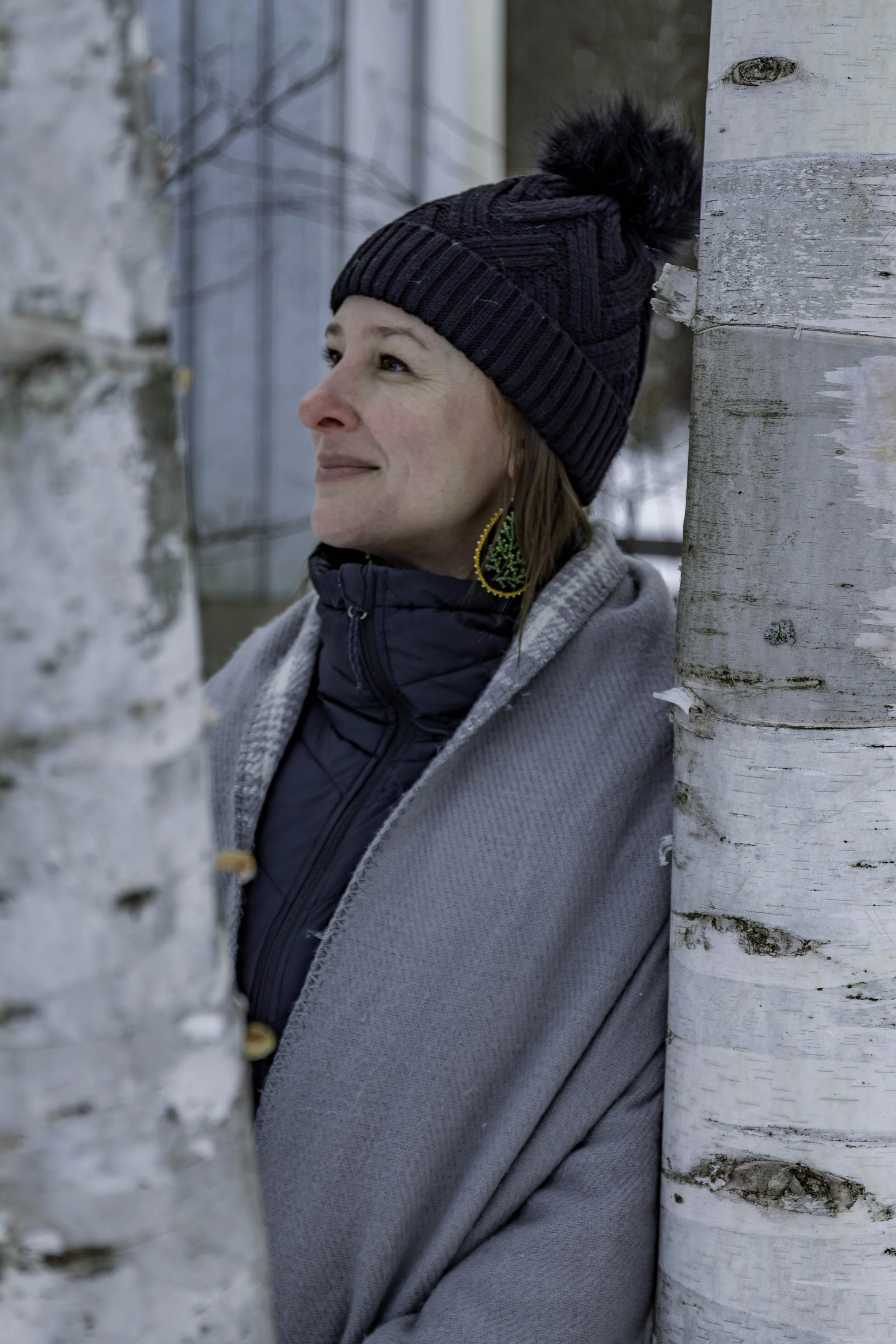 Cristin is wearing a dark knit hat with a pompom, a gray coat, and a blue jacket underneath, standing between two white birch trees outside during winter, smiling softly with her eyes closed.