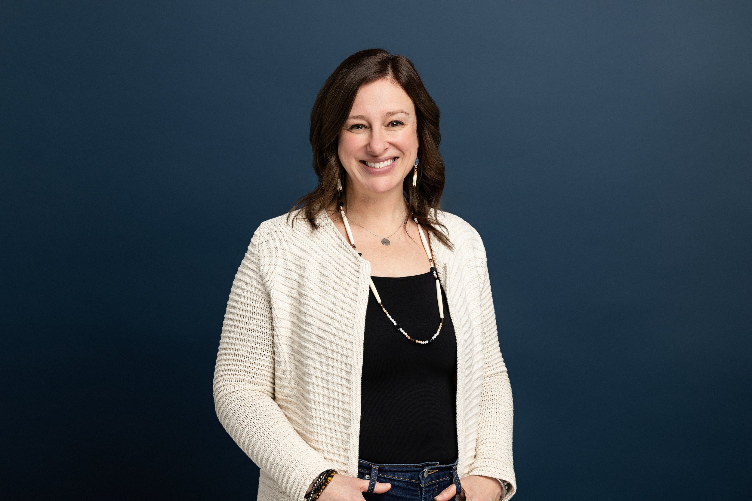 Cristin is wearing a cream cardigan over a black top, with beaded jewelry, standing against a blue background.