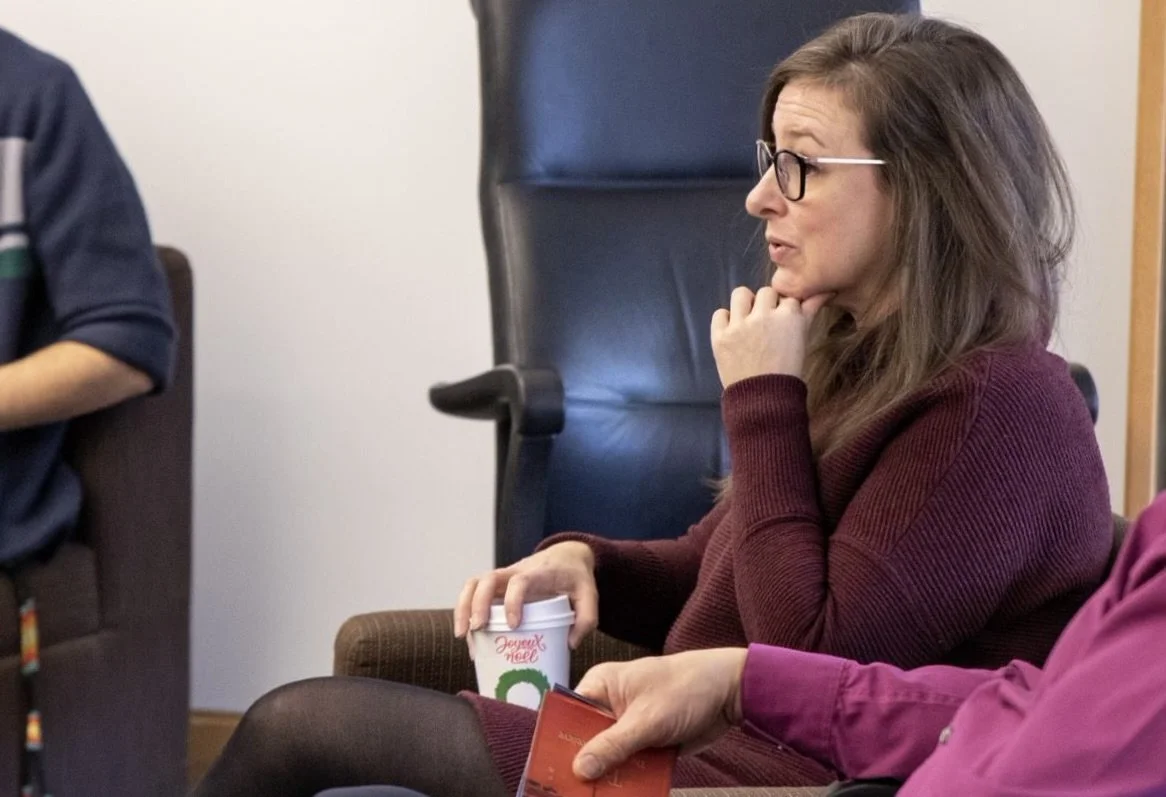 Cristin with glasses and long brown hair, wearing a maroon sweater, sitting in a chair during a meeting or discussion, holding a cup of coffee with her chin resting on her hand.