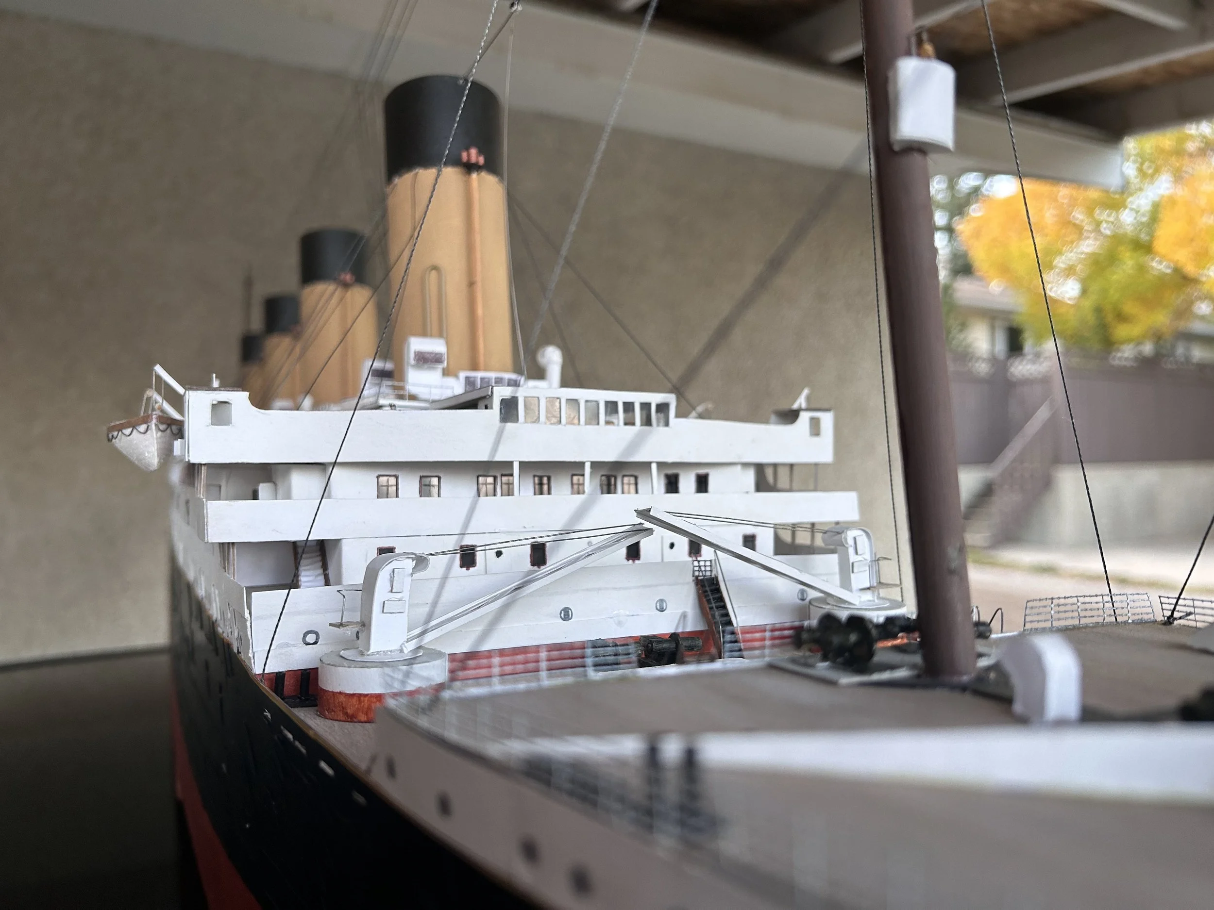 Model of a ship with three smokestacks, viewed from a low angle with a background of a porch ceiling and autumn trees.