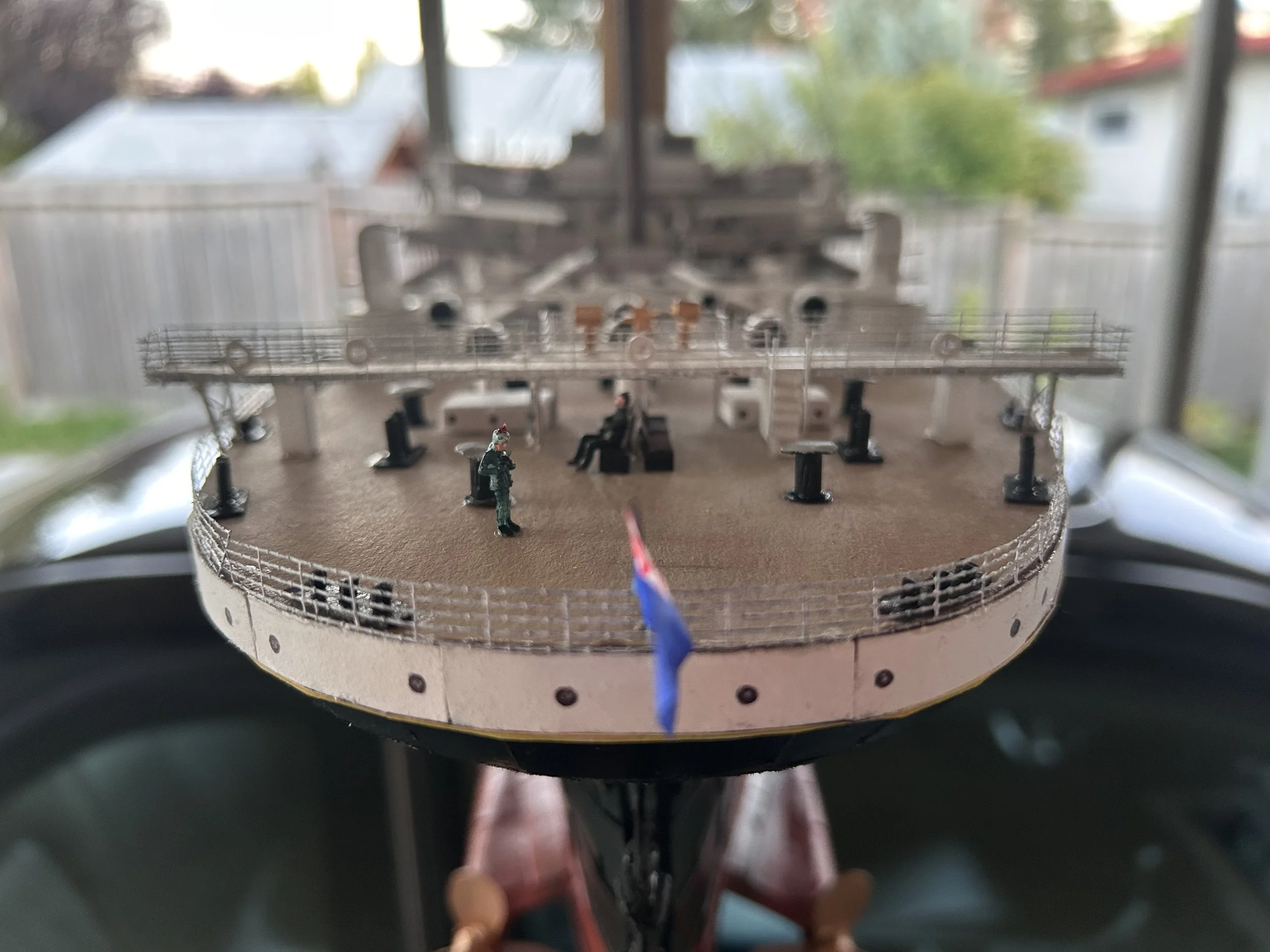 Close-up of a detailed model ship deck with miniature figures, railings, and equipment, with a blurred outdoor background.