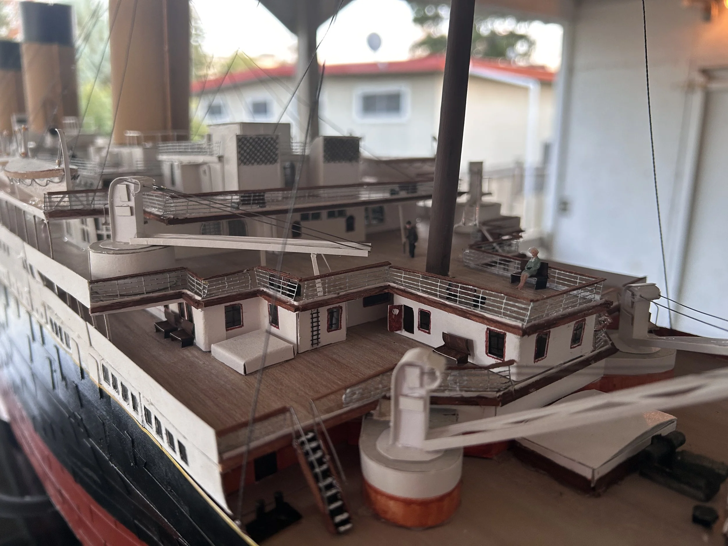 Model of a ship's deck with small buildings, railings, and a miniature figure of a person sitting on a bench, with wires and support structures overhead.