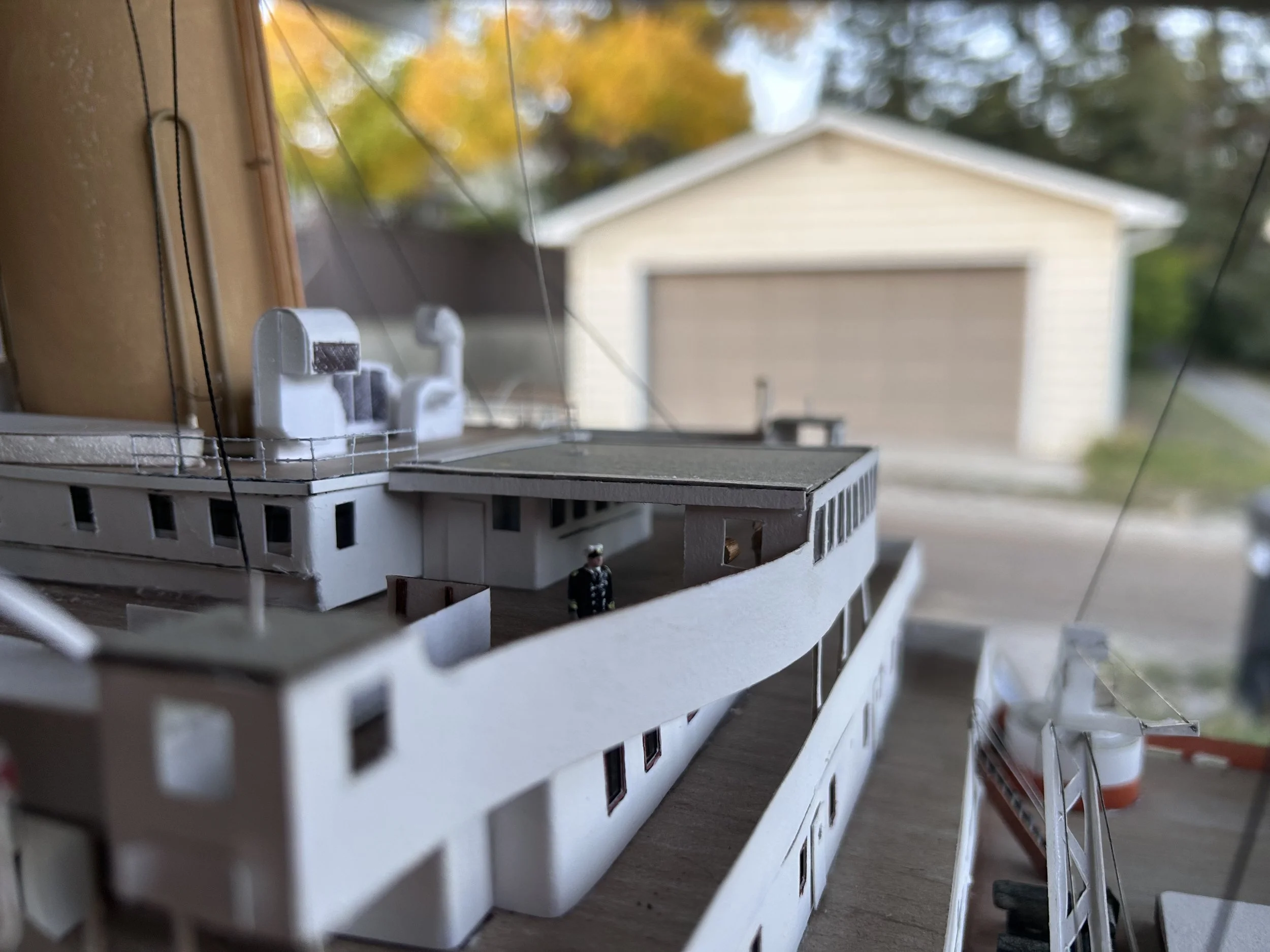 Close-up of a detailed architectural model of a building, with miniature figures, set indoors near a window, with a garage and trees visible outside.