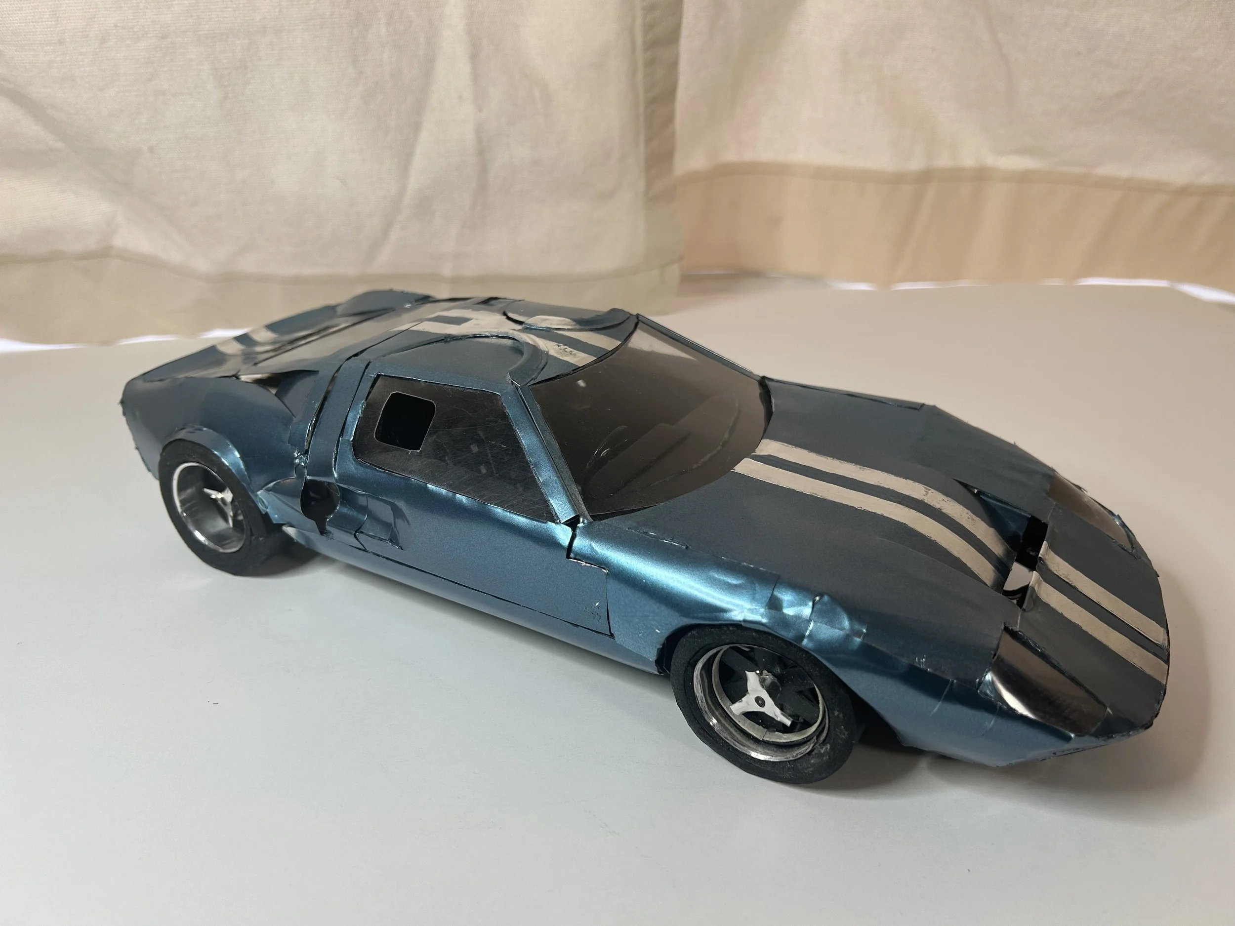 A damaged toy model of a blue sports car with white racing stripes on the hood, placed on a white surface with a beige curtain backdrop.