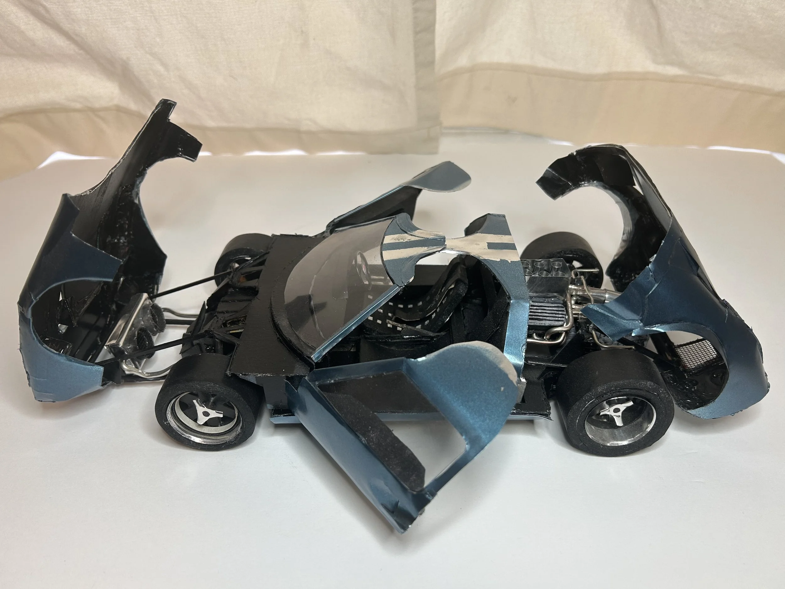 Model of a crashed sports car with broken body panels and exposed internal components, displayed on a white surface.