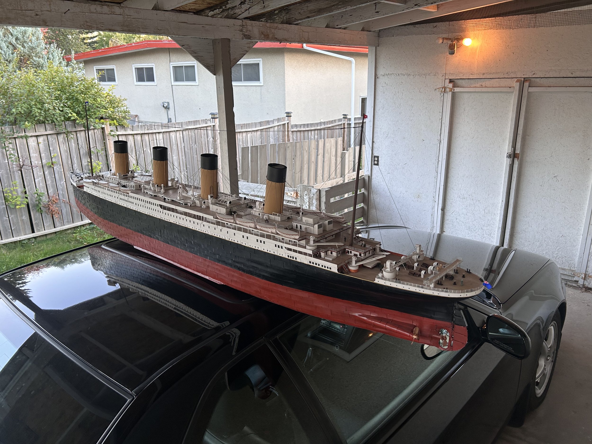 Model of the Titanic ship placed on the roof of a black car in a garage with a wooden fence and houses in the background.