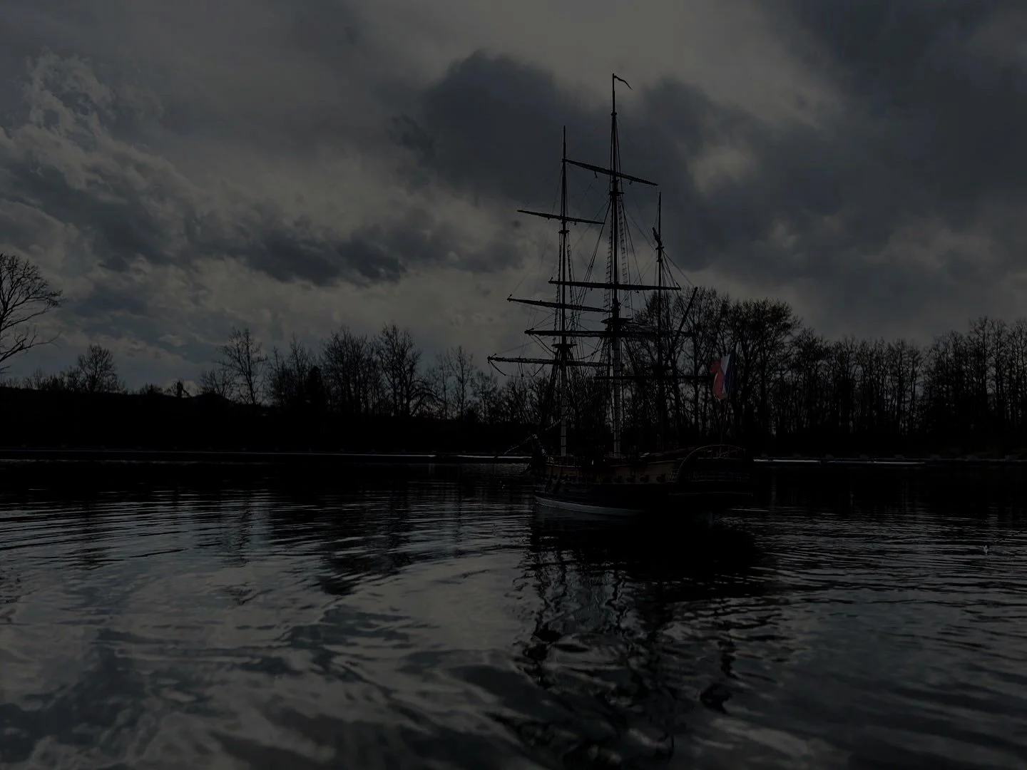A dark, moody scene of a tall sailing ship with multiple masts and rigging, docked on a calm body of water under a cloudy sky with leafless trees in the background.