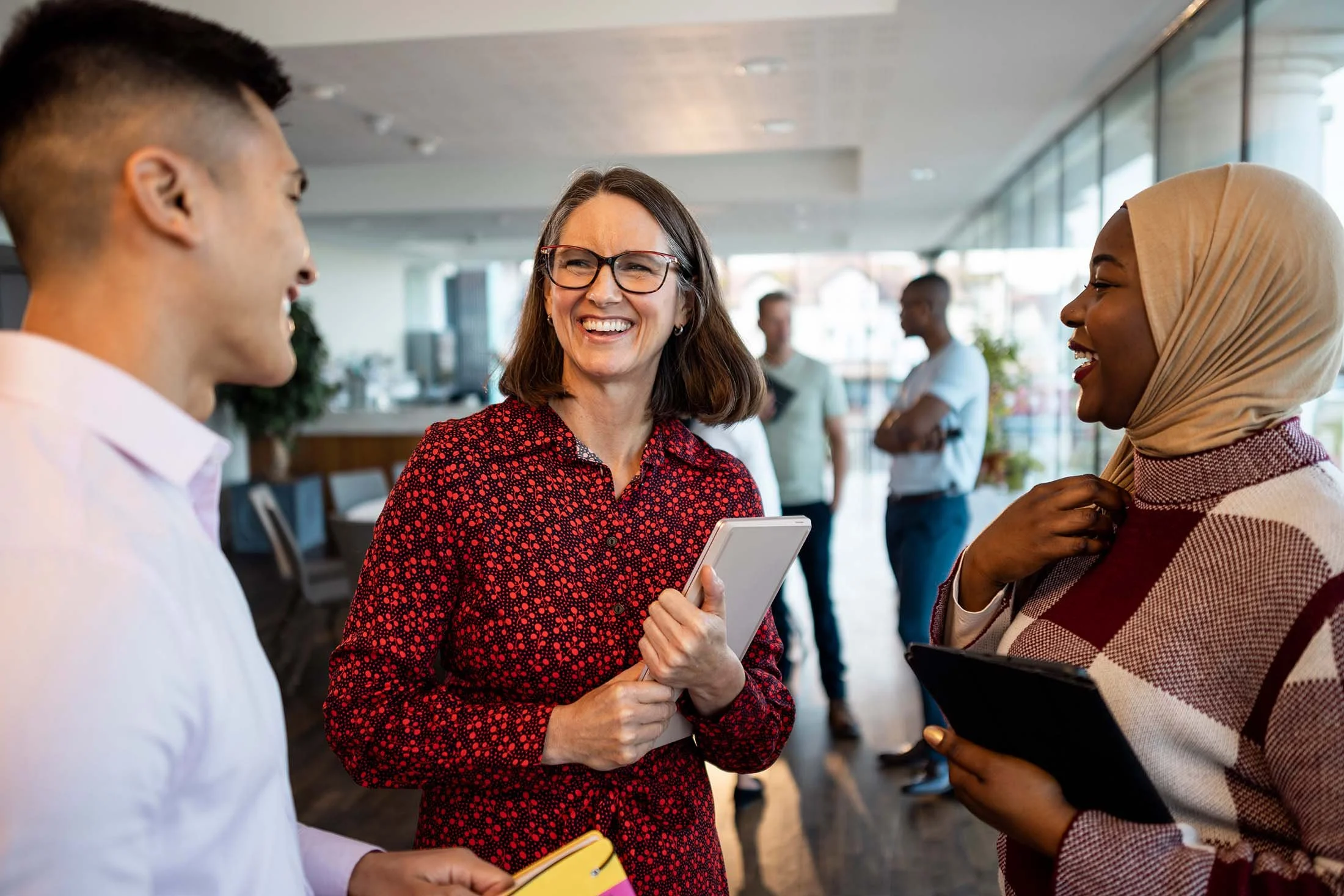 Three diverse people engaged in conversation at a social or professional event, with a woman in glasses holding a tablet and smiling.