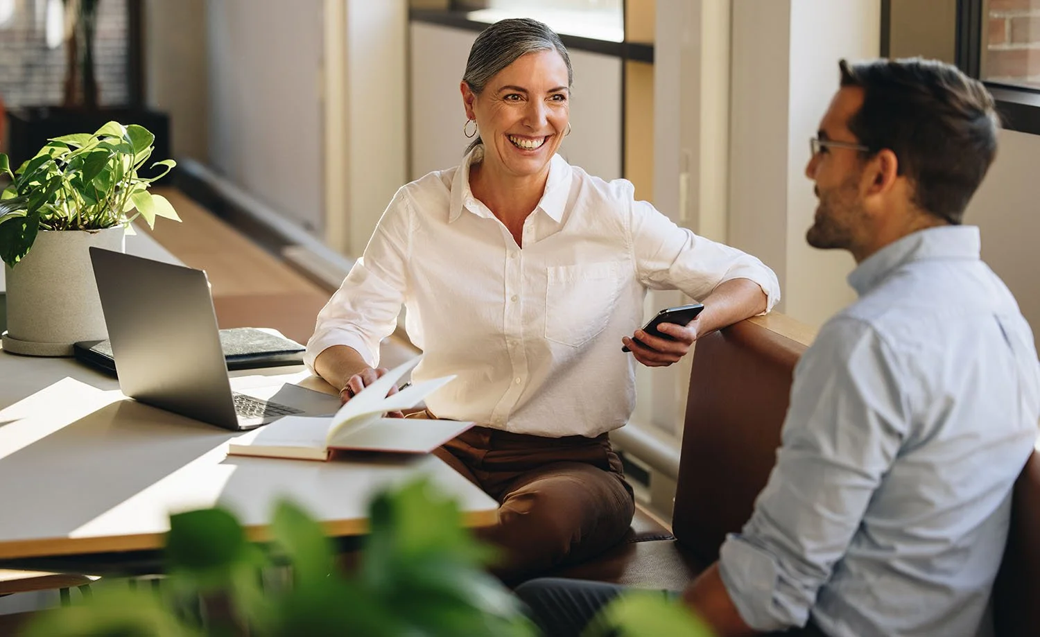 A woman and man sitting at a desk in an office, engaged in conversation, smiling. The woman is holding a smartphone, and there is a laptop, open books, and a plant on the desk.