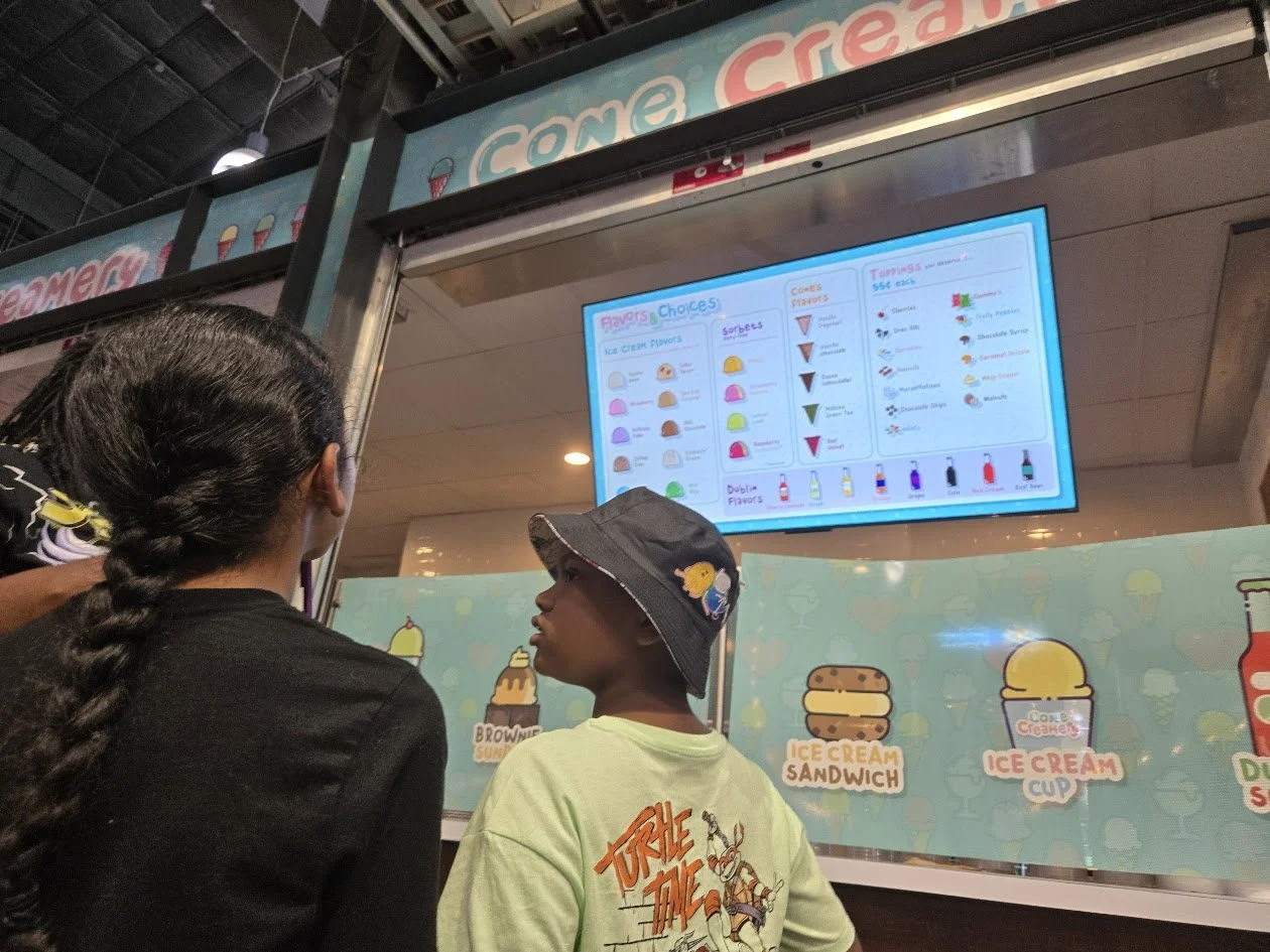 Two people standing at an ice cream shop menu, looking at the menu board which displays various ice cream flavors, sorbets, toppings, and prices during a Beulah's Bunch field trip.