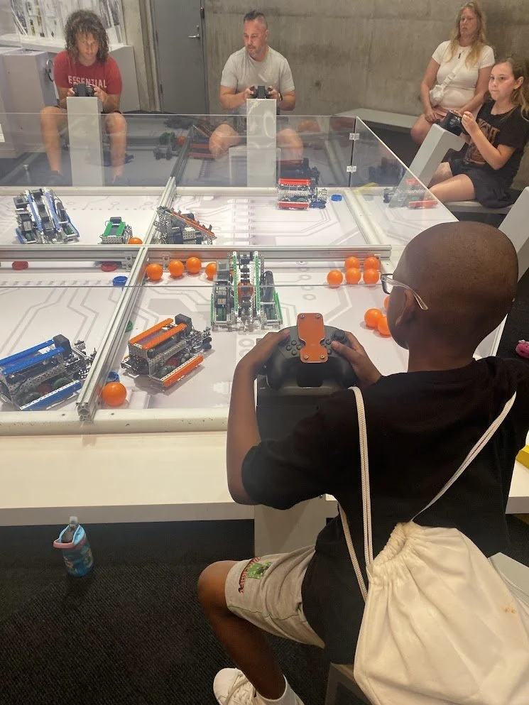 Children playing a robotics game with orange balls at a table enclosed by transparent barriers during a field trip to the planetarium.
