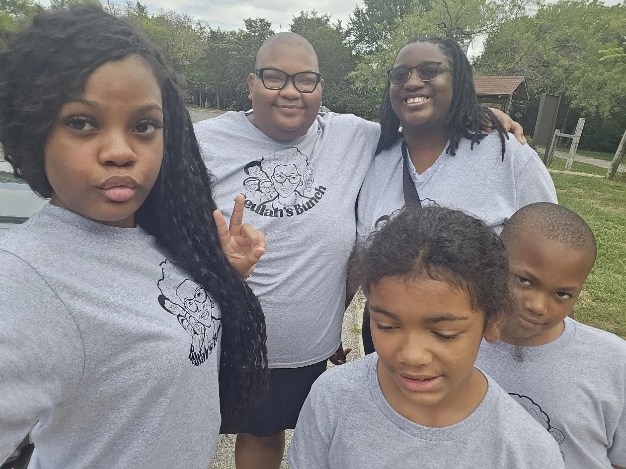 Group of six people outdoors, wearing matching gray T-shirts with a cartoon graphic and 'Beulah's Bunch merch tees, smiling and posing for a selfie.