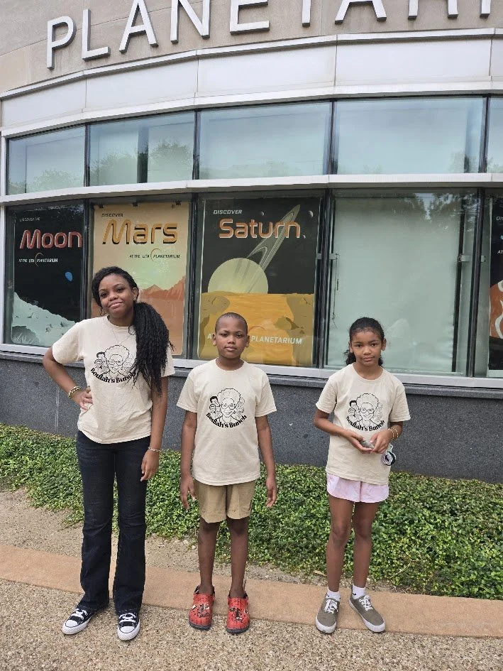 Three children standing outside a planetarium with posters of Mars and Saturn behind them, wearing matching beige t-shirts with a black logo, and smiling during a field trip with Beulah's Bunch Camp organizers
.