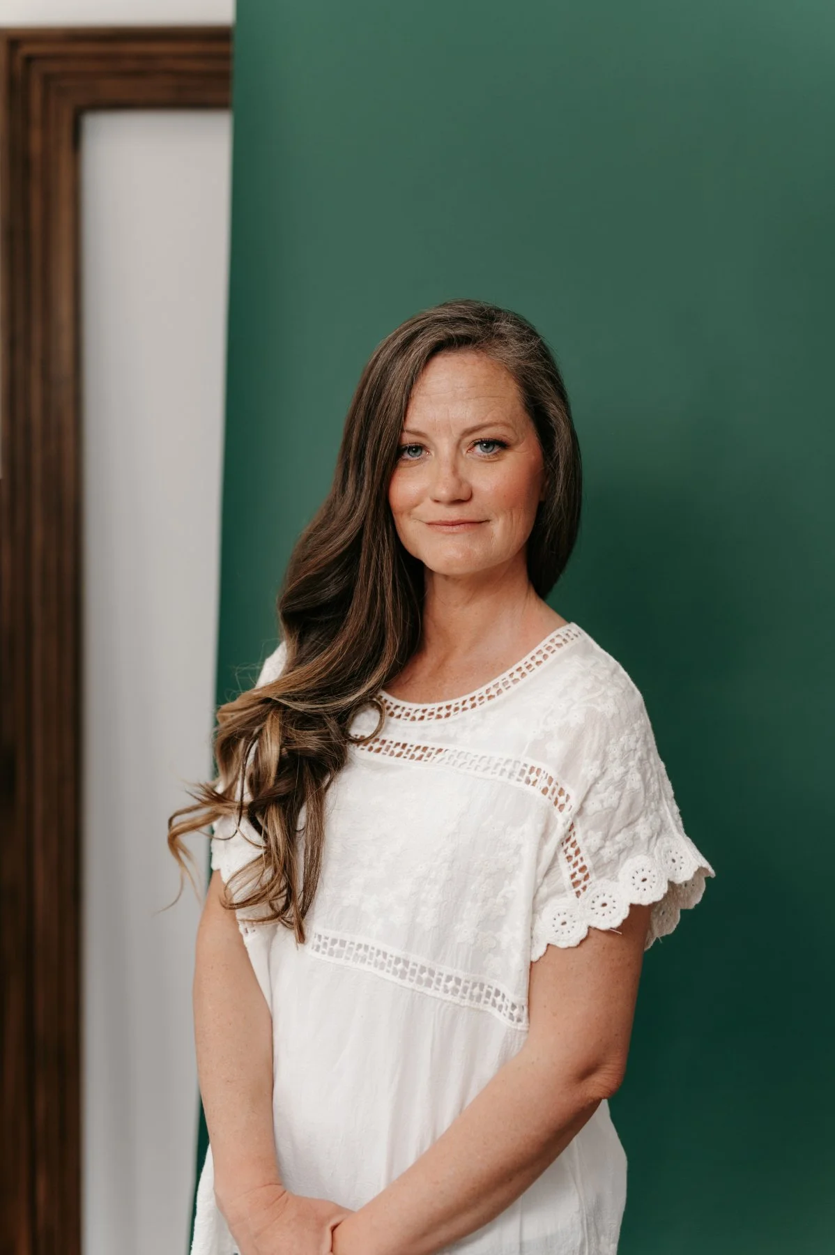 A woman with long, wavy brown hair standing in front of a green background, wearing a white embroidered blouse, looking at the camera with a slight smile.