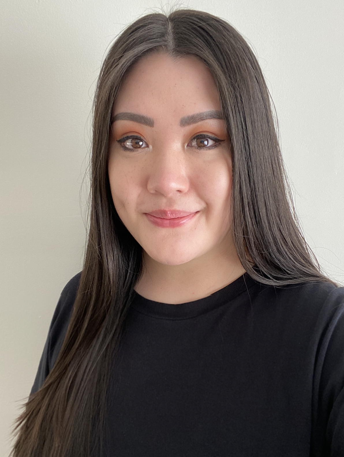 A young woman with long dark hair, light skin, and makeup, wearing a black shirt, smiling against a neutral background.