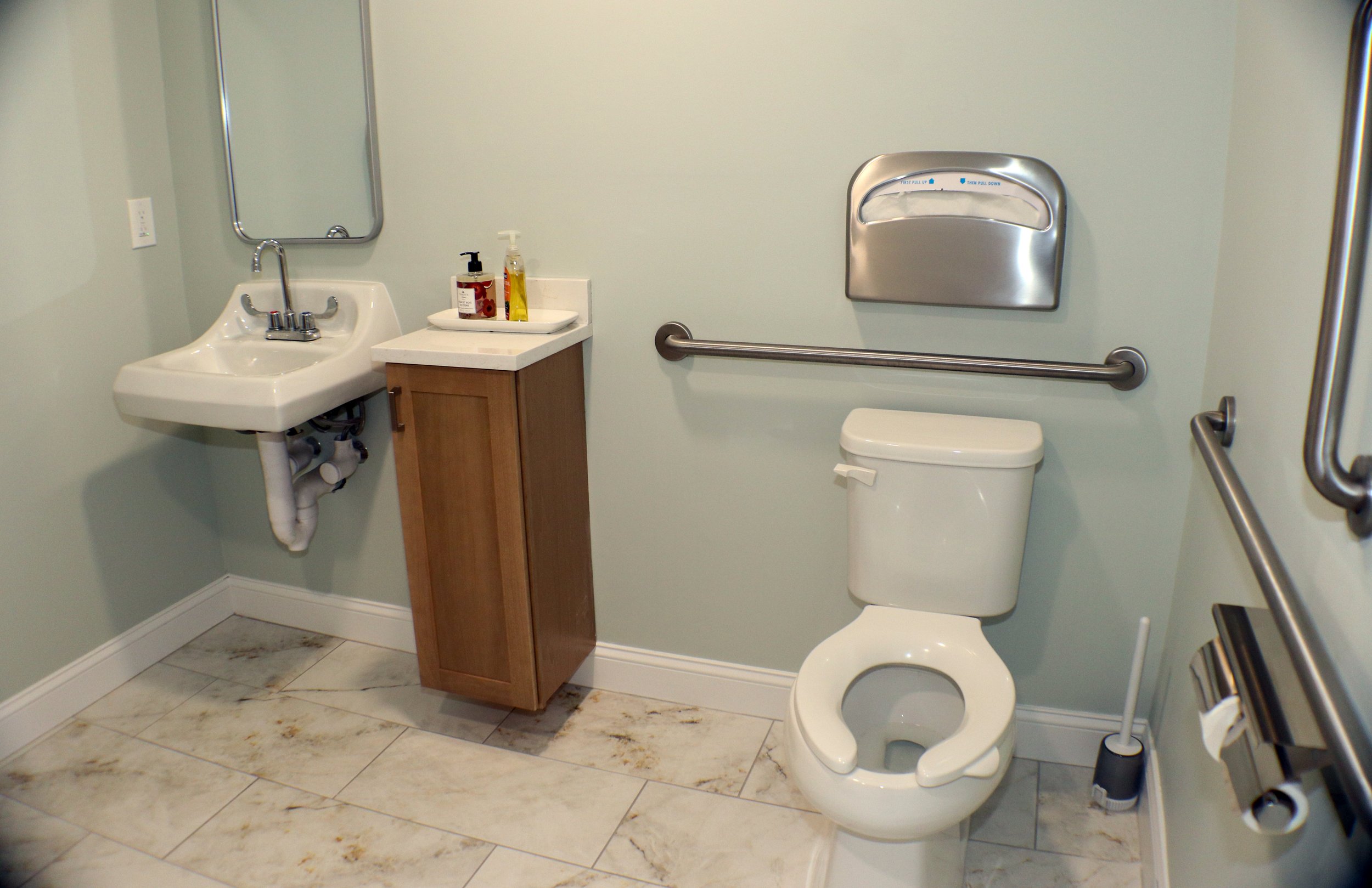 A bathroom with a small sink, a mirror, a wooden cabinet with soap and hand sanitizer, a toilet with a support rail, a wall-mounted paper towel dispenser, a toilet brush, and handrails on the walls.