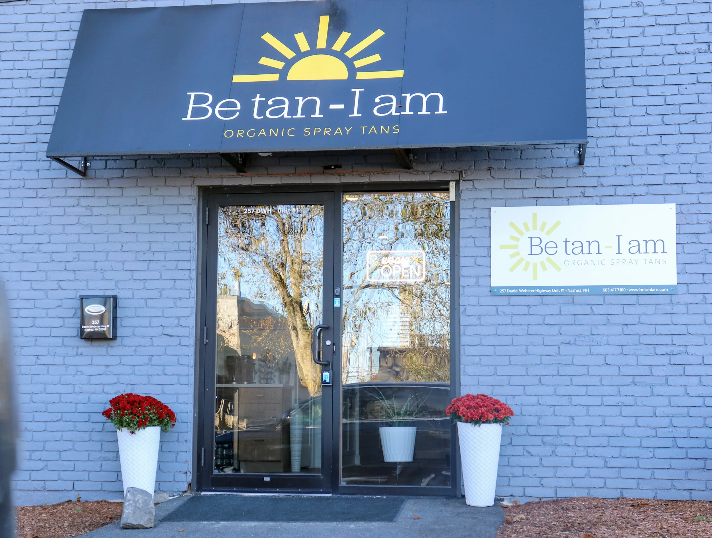 The storefront of Be tan-I am, an organic spray tan business, features a large sign with a yellow sunburst logo and the business name. The entrance has a glass door with an 'Open' sign. There are two tall white planters with red flowers on either sid