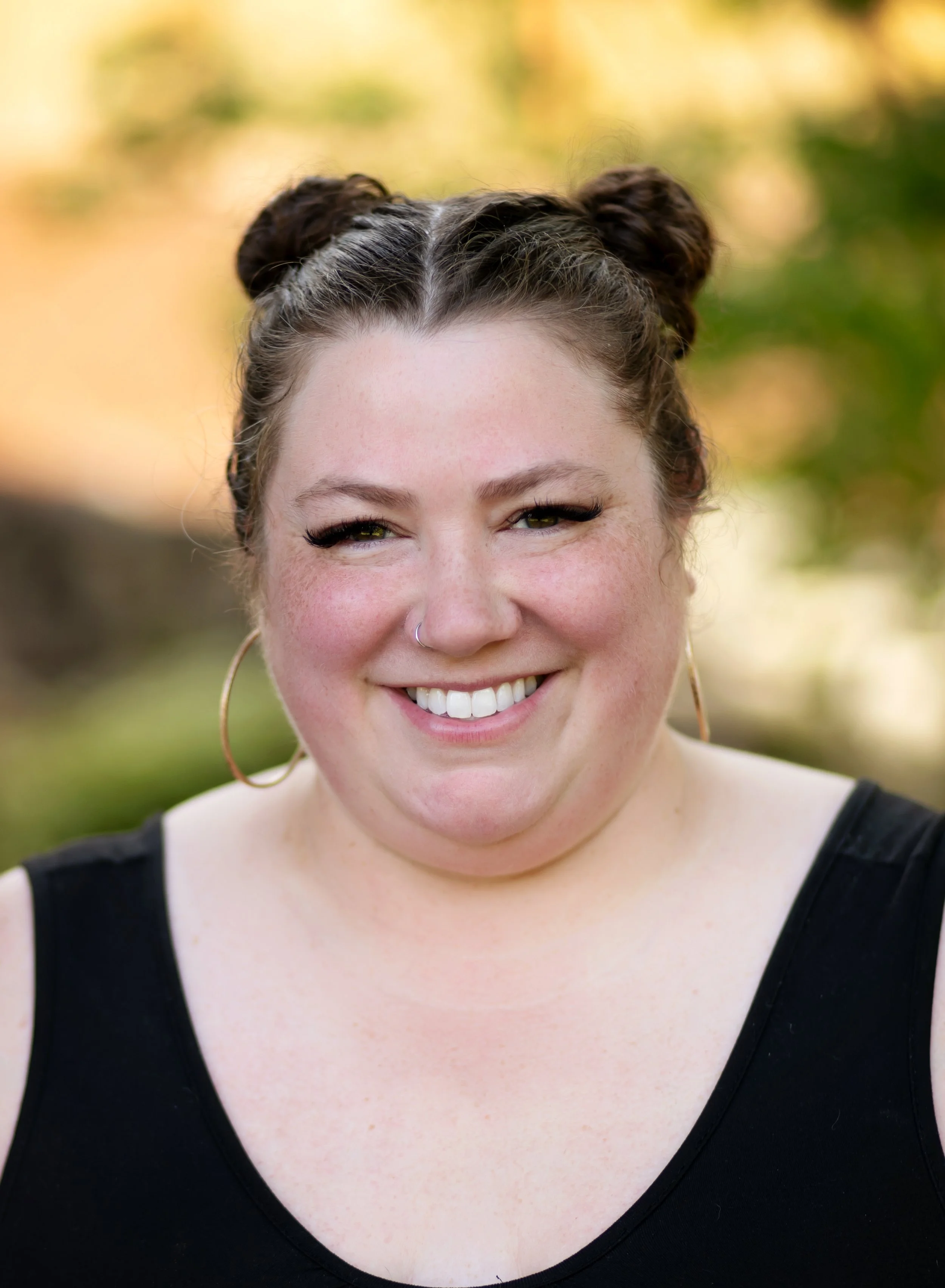 A young woman with light skin smiling, wearing large gold hoop earrings and a black top, outdoors with a blurred yellow and green background.