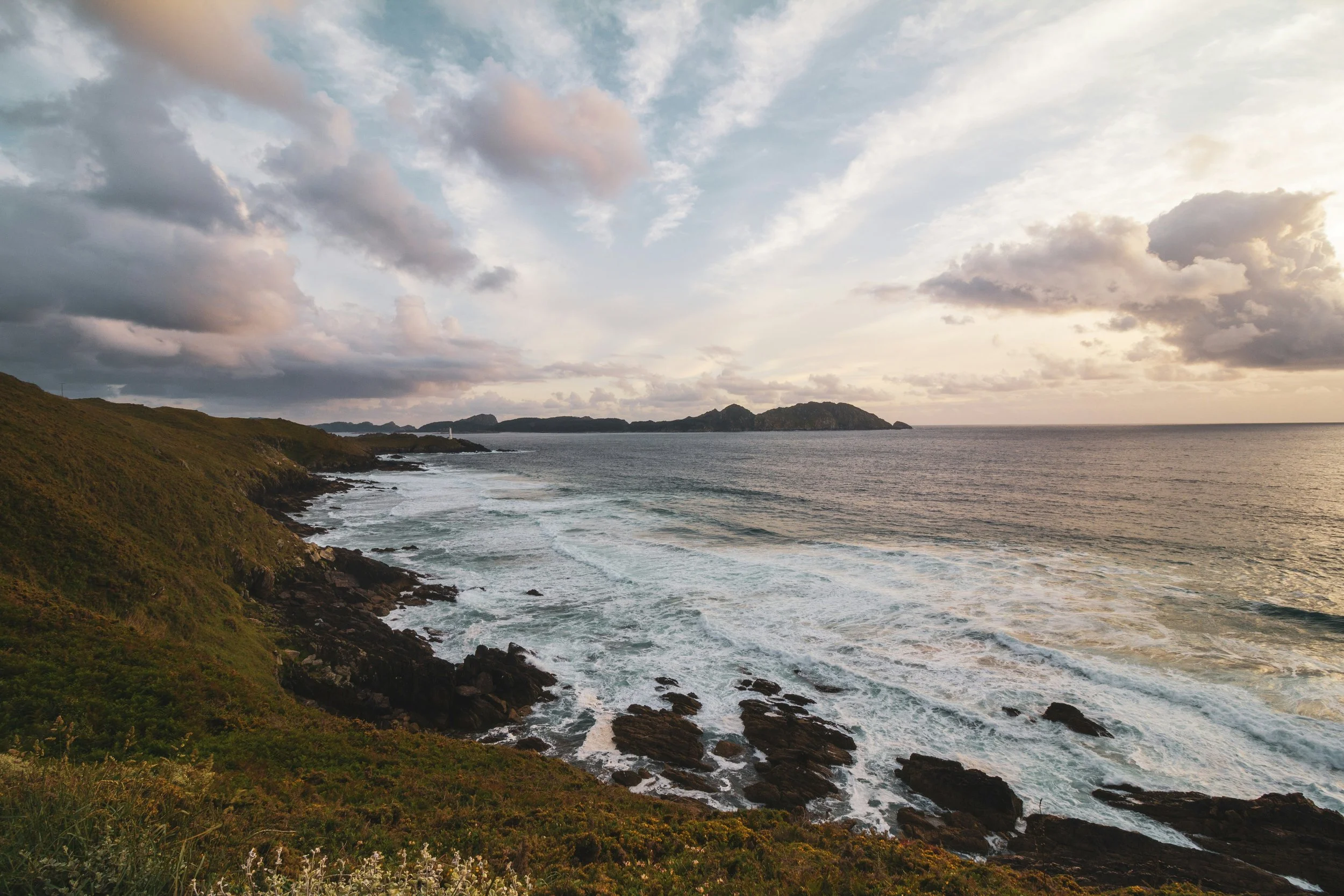 A coastal scene with green hills on the left, rocky shoreline, and ocean waves under a sky with scattered clouds at sunset.