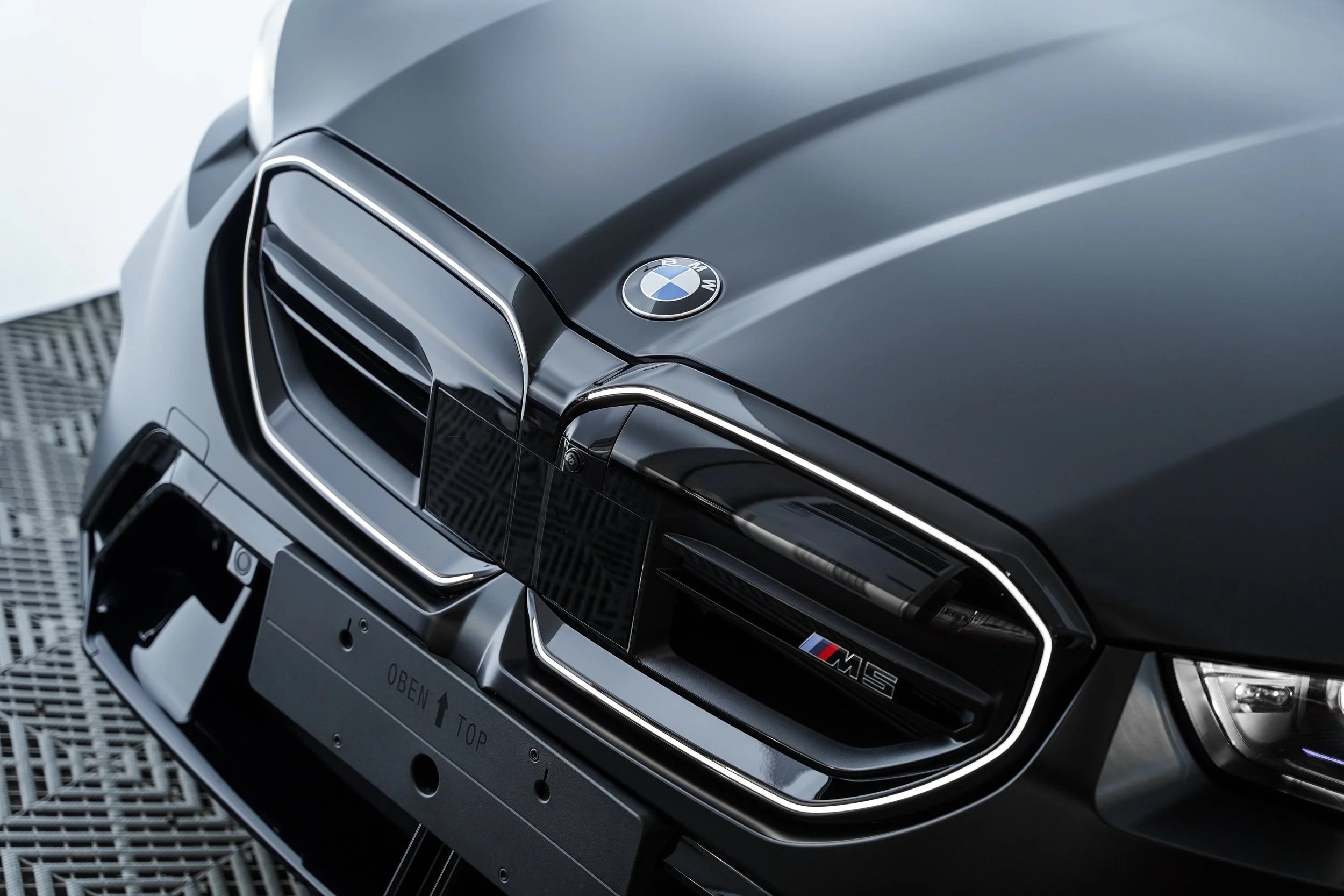 Close-up of a matte gray BMW M5 front grille, with the BMW logo on the hood and M5 badge, man-made grille design, and part of the front bumper visible.