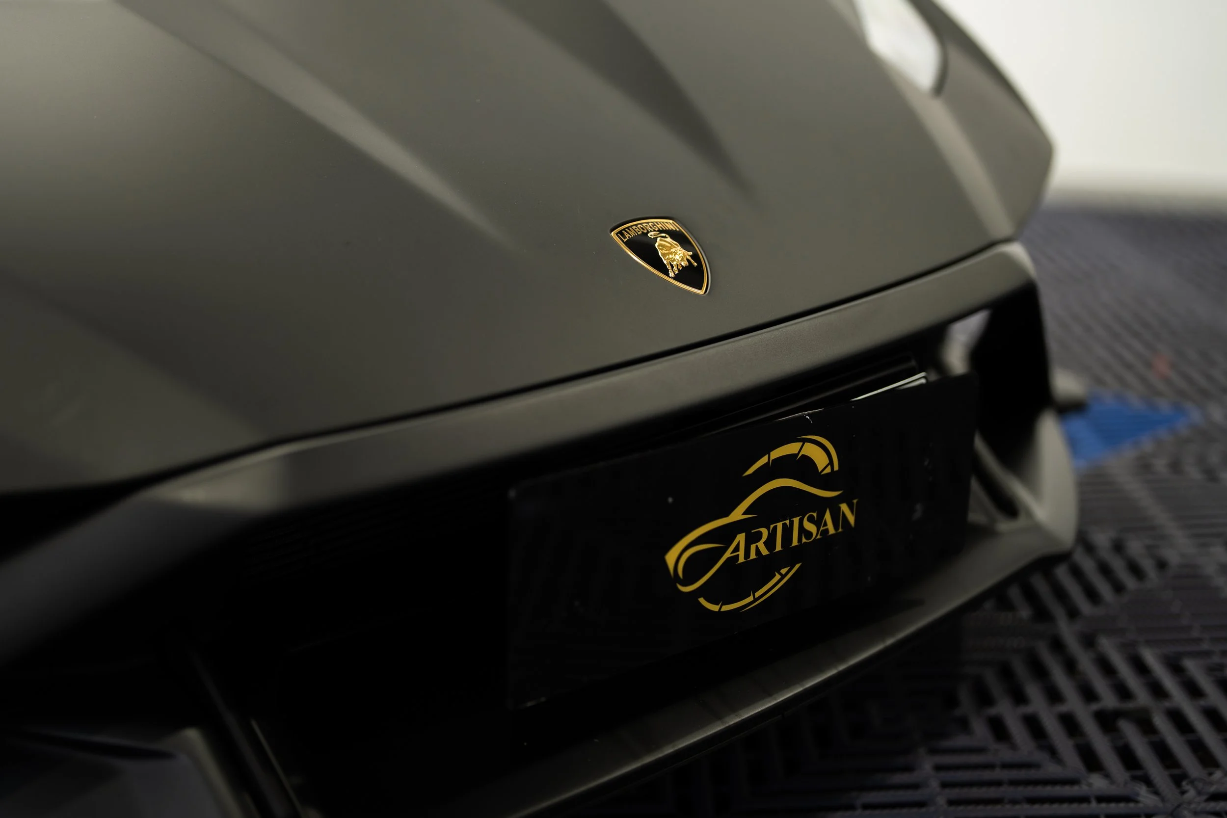 Close-up of a black Lamborghini car badge on a gray vehicle and a black plate with gold writing that says 'Artisan'.